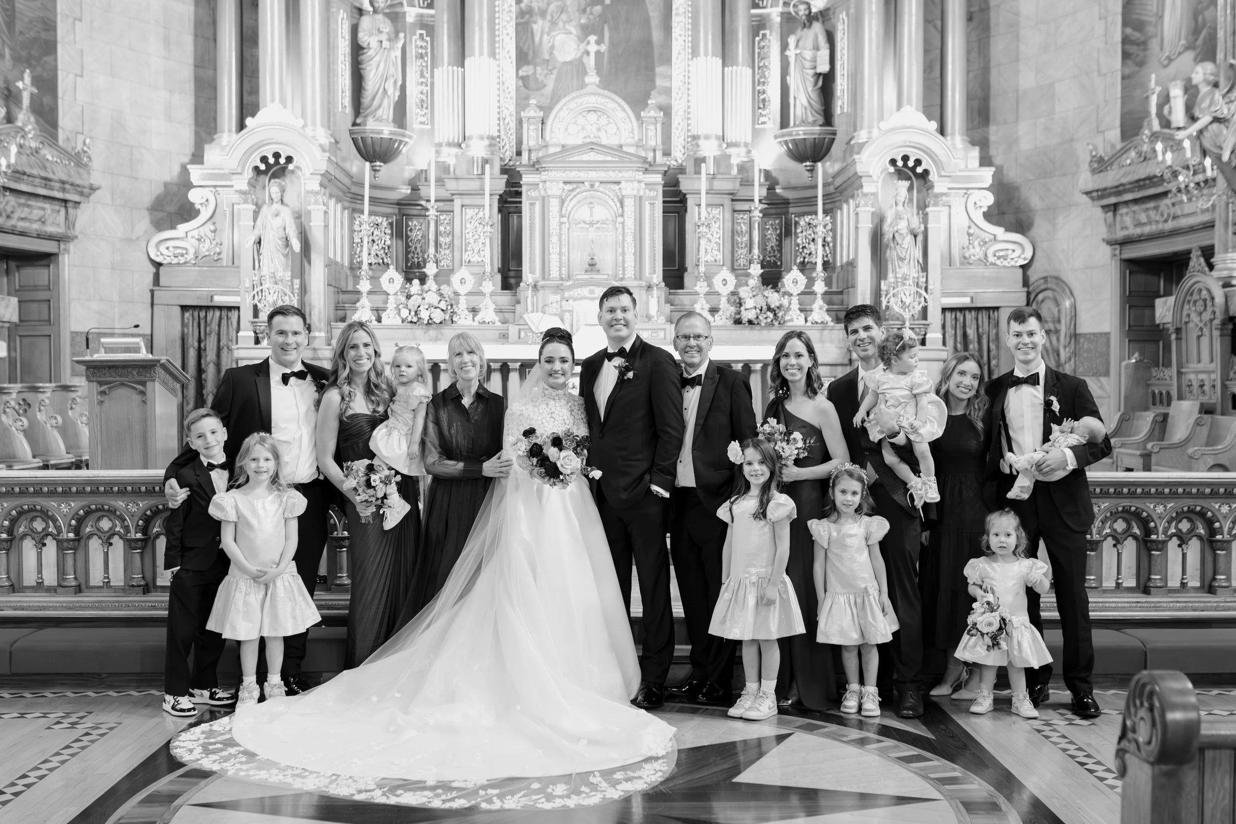 A wedding group photo inside a church, featuring the bride and groom at the center surrounded by children and adults, all dressed in formal attire.