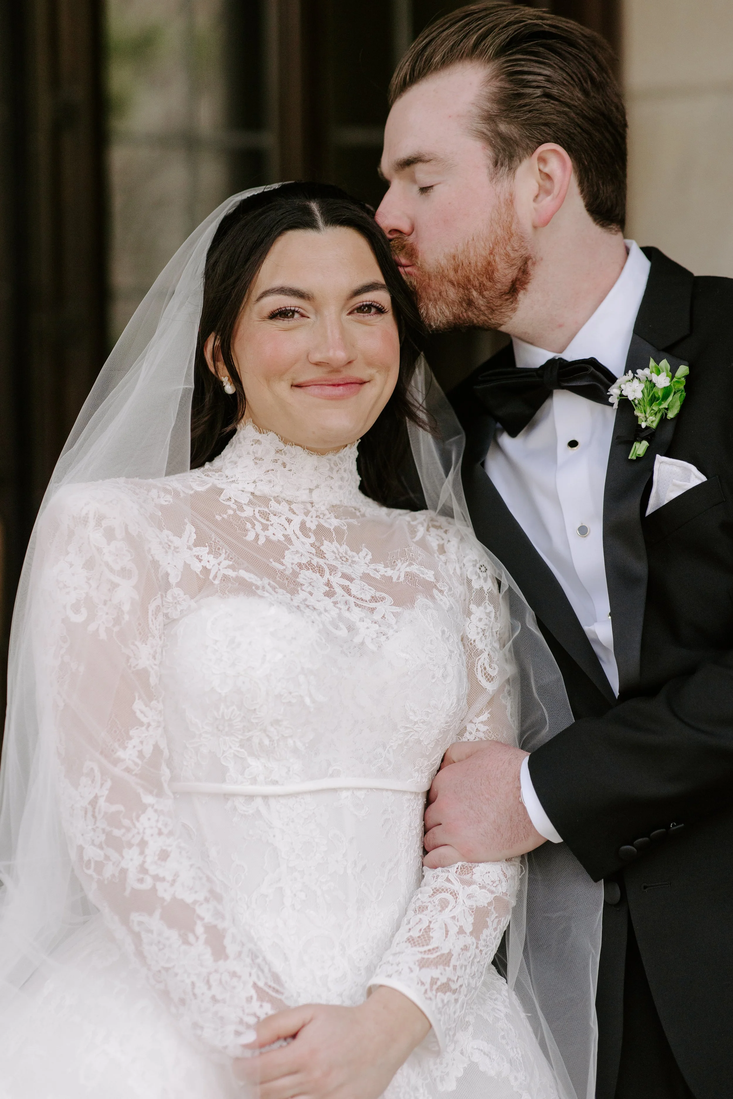 A bride and groom on their wedding day, with the groom kissing the bride on her temple. The bride is smiling softly, wearing a lace wedding gown and veil, while the groom is dressed in a black tuxedo with a boutonniere.