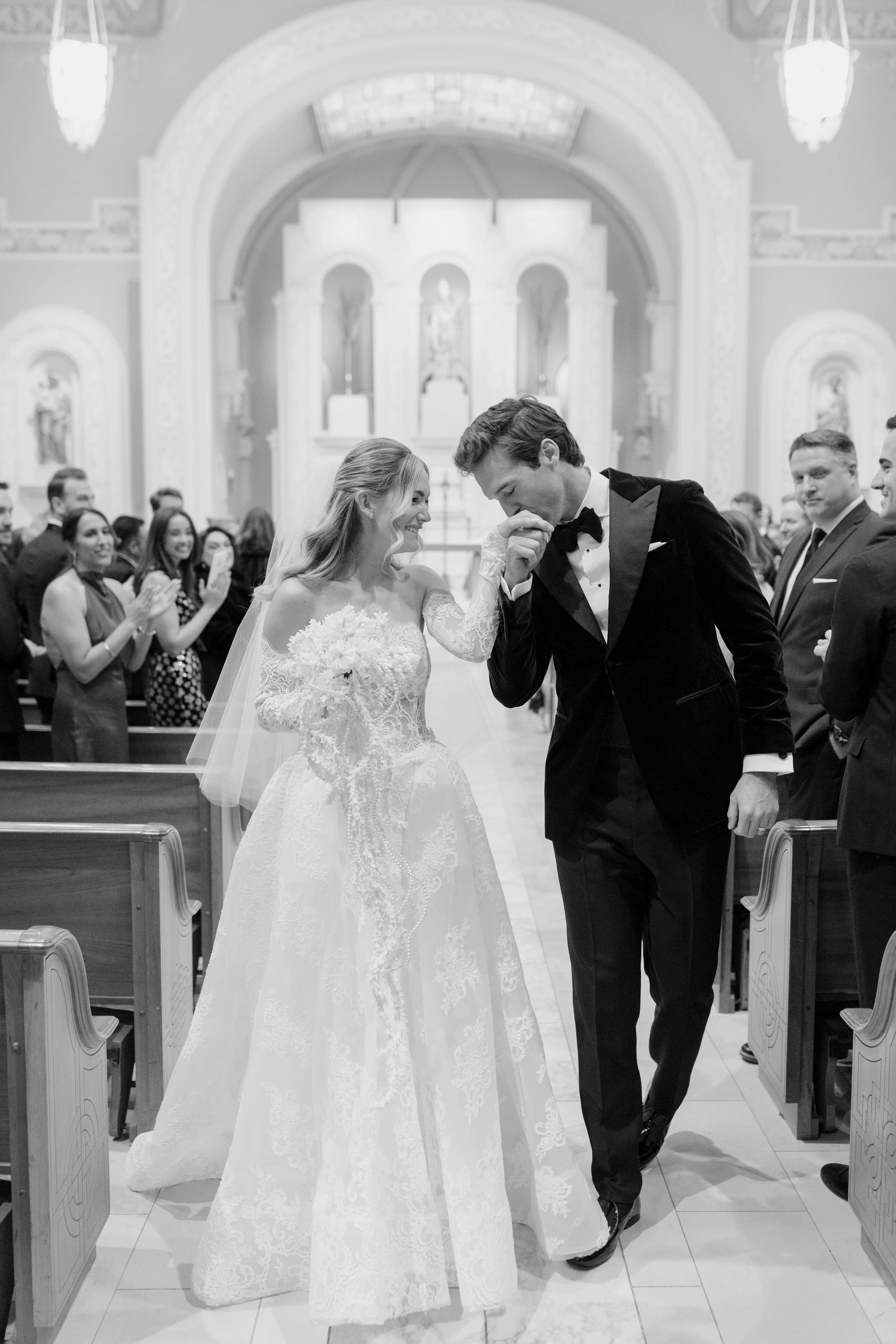 A bride and groom kissing in a church during their wedding ceremony. The bride is wearing a lace wedding gown and veil, and the groom is dressed in a tuxedo. Guests are clapping and smiling in the background.