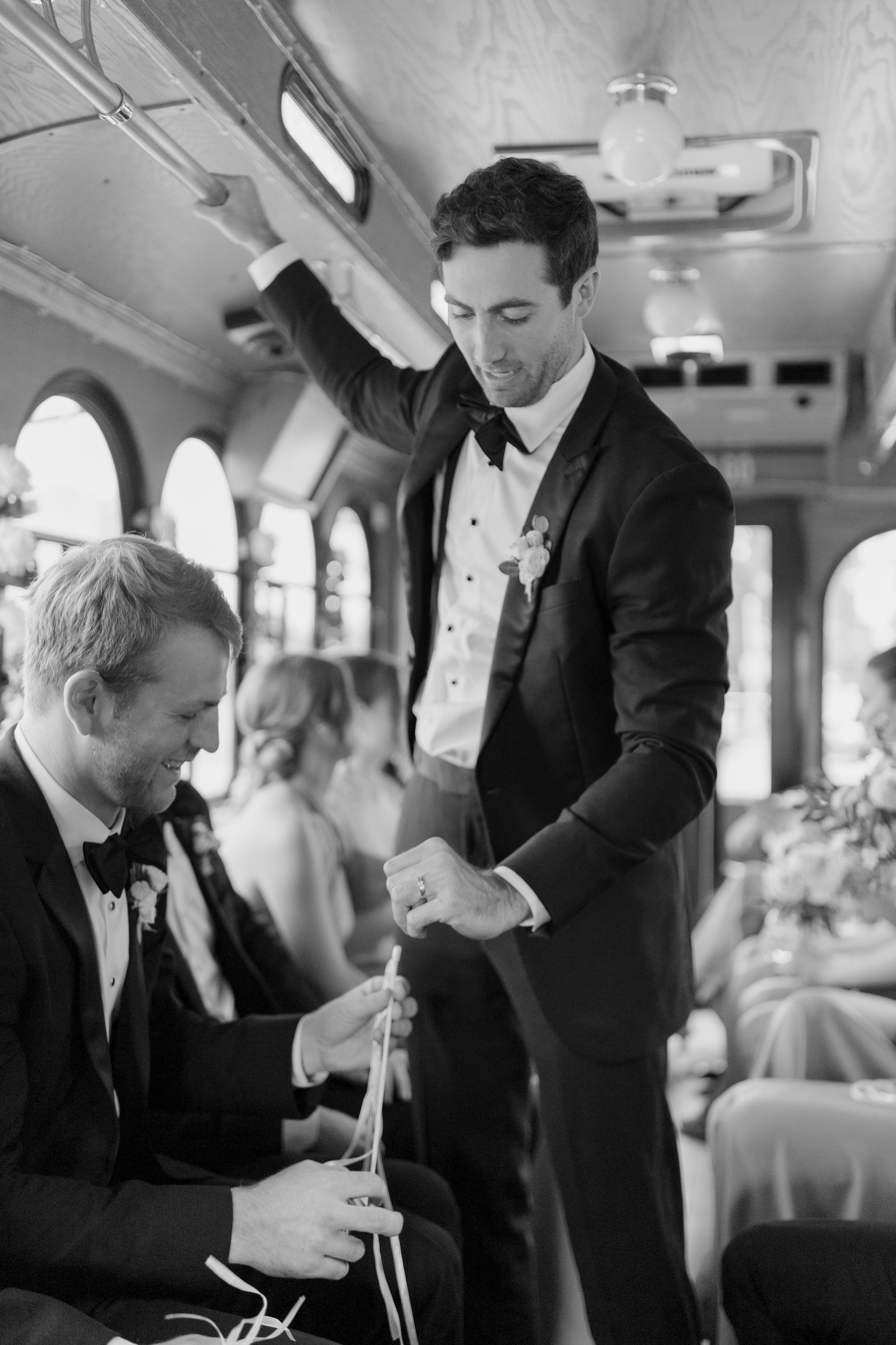 A man in a tuxedo with a bow tie and boutonniere is standing inside a bus, leaning on a handrail, with other seated passengers, engaged in a conversation or activity at a gathering or wedding event.