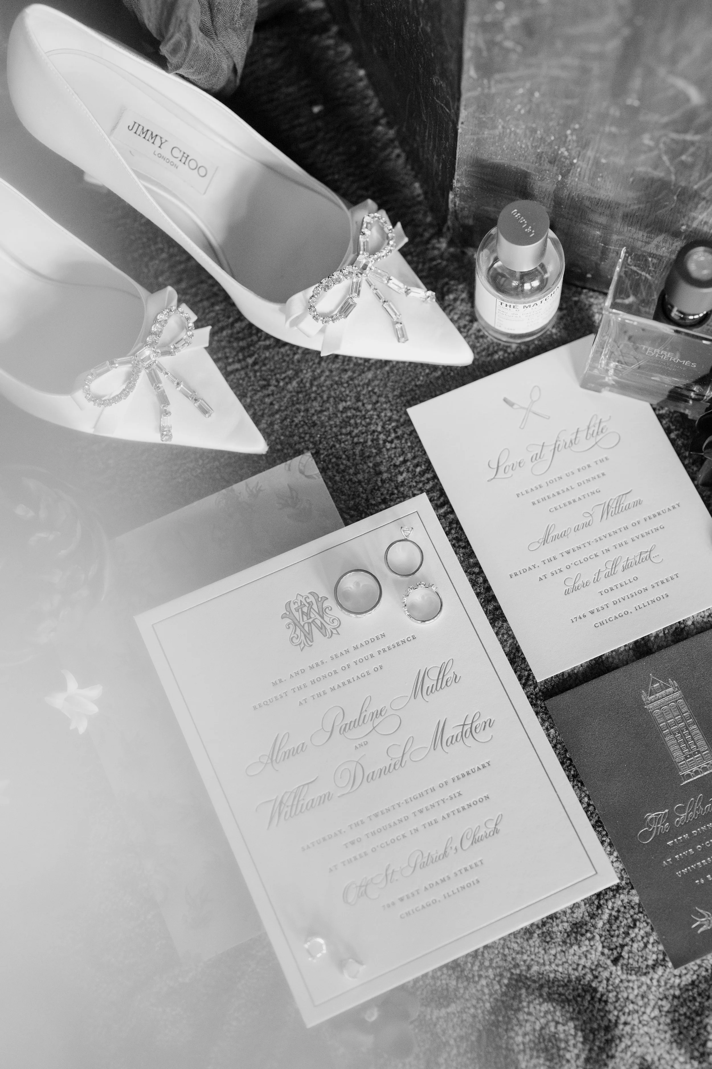 Black and white photo showing wedding invitations and accessories on a table. The invitations feature elegant cursive text and monograms, with rings and jewelry nearby. A pair of white shoes with bows and rhinestone embellishments are also visible.