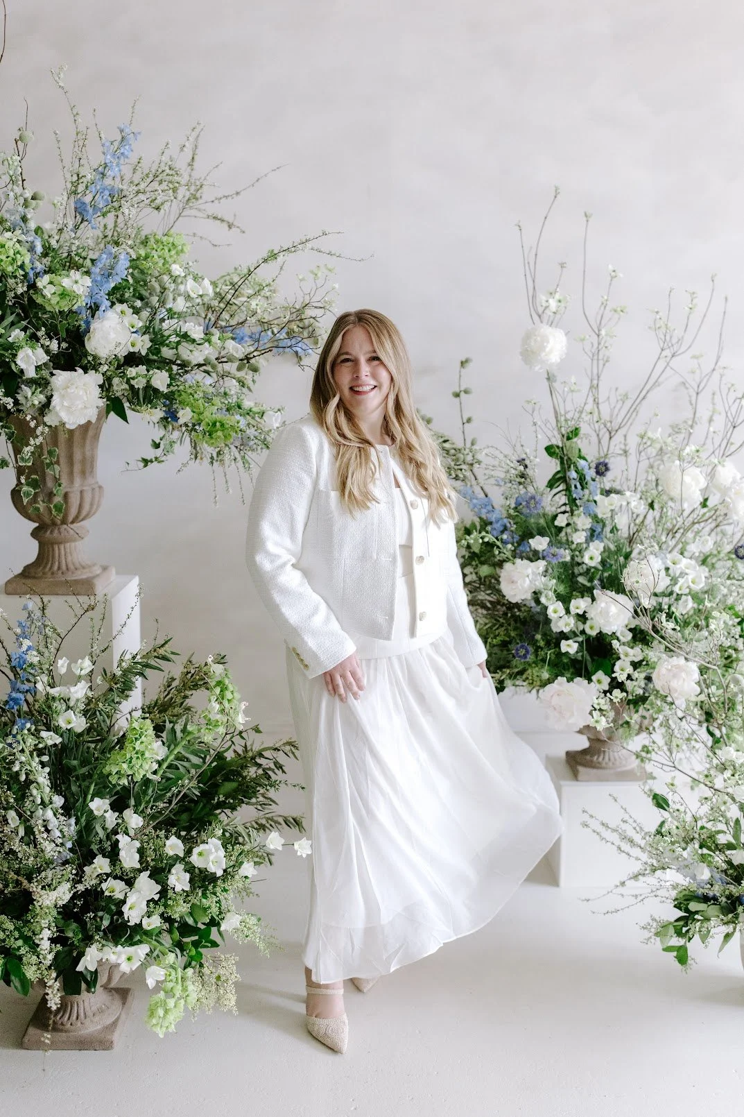 A smiling woman in a white dress and jacket standing near floral arrangements with white, green, and blue flowers on white pedestals and background.