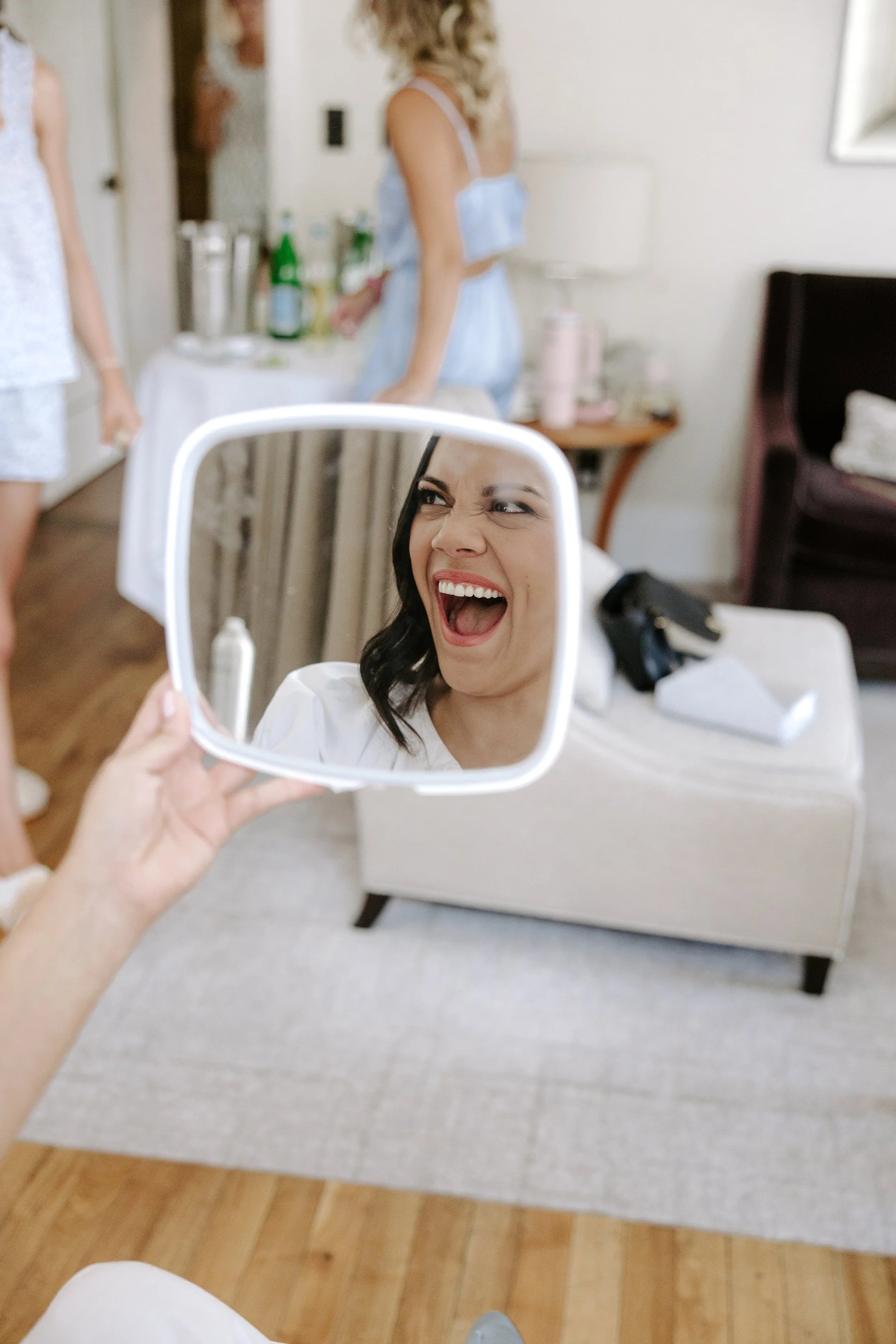 A woman with dark hair smiling and making a silly face while looking into a handheld mirror.