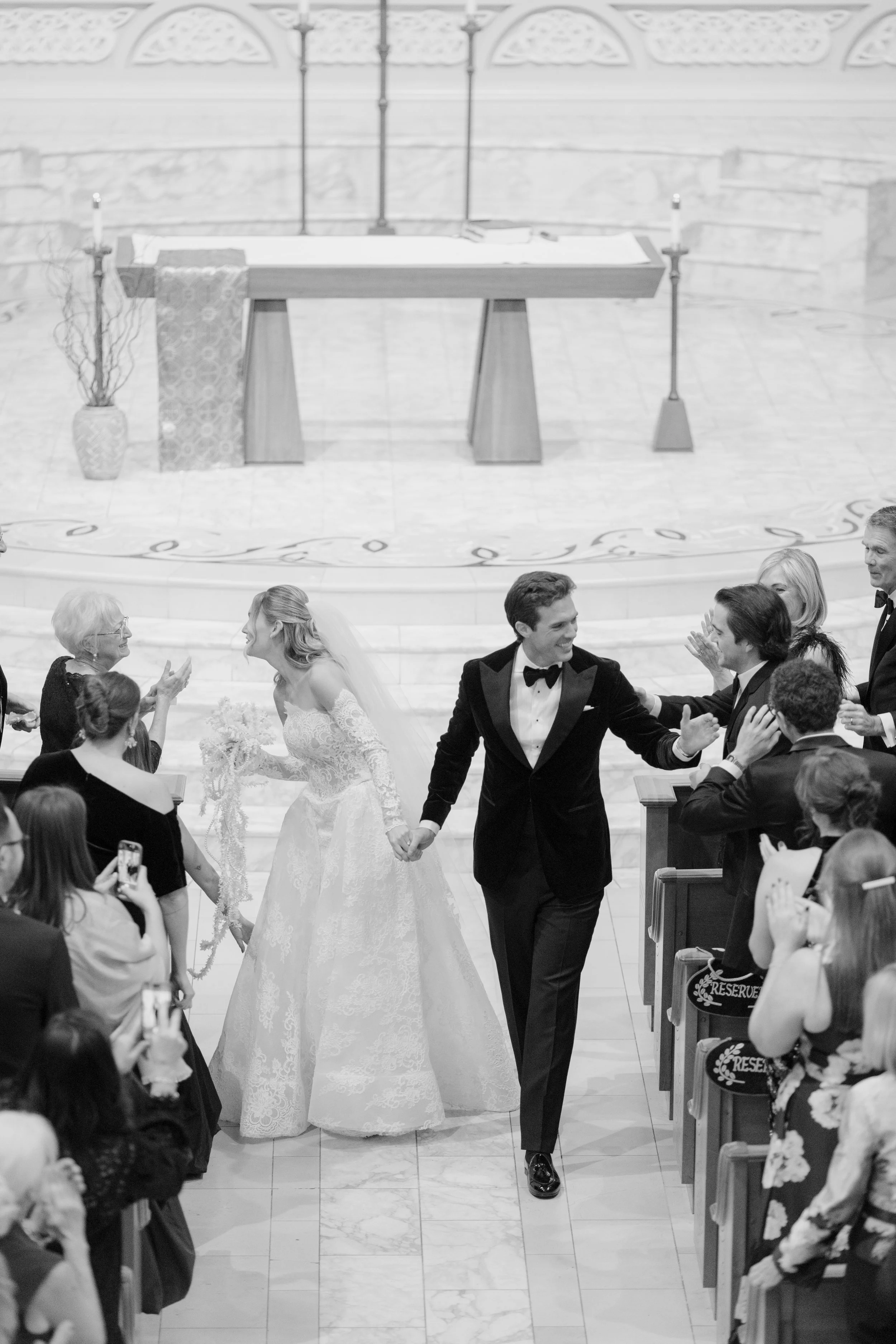 A bride and groom are holding hands and walking down the aisle in a church during their wedding ceremony. Guests on both sides are clapping and taking photos. The church has a high ceiling and a small table in the background.