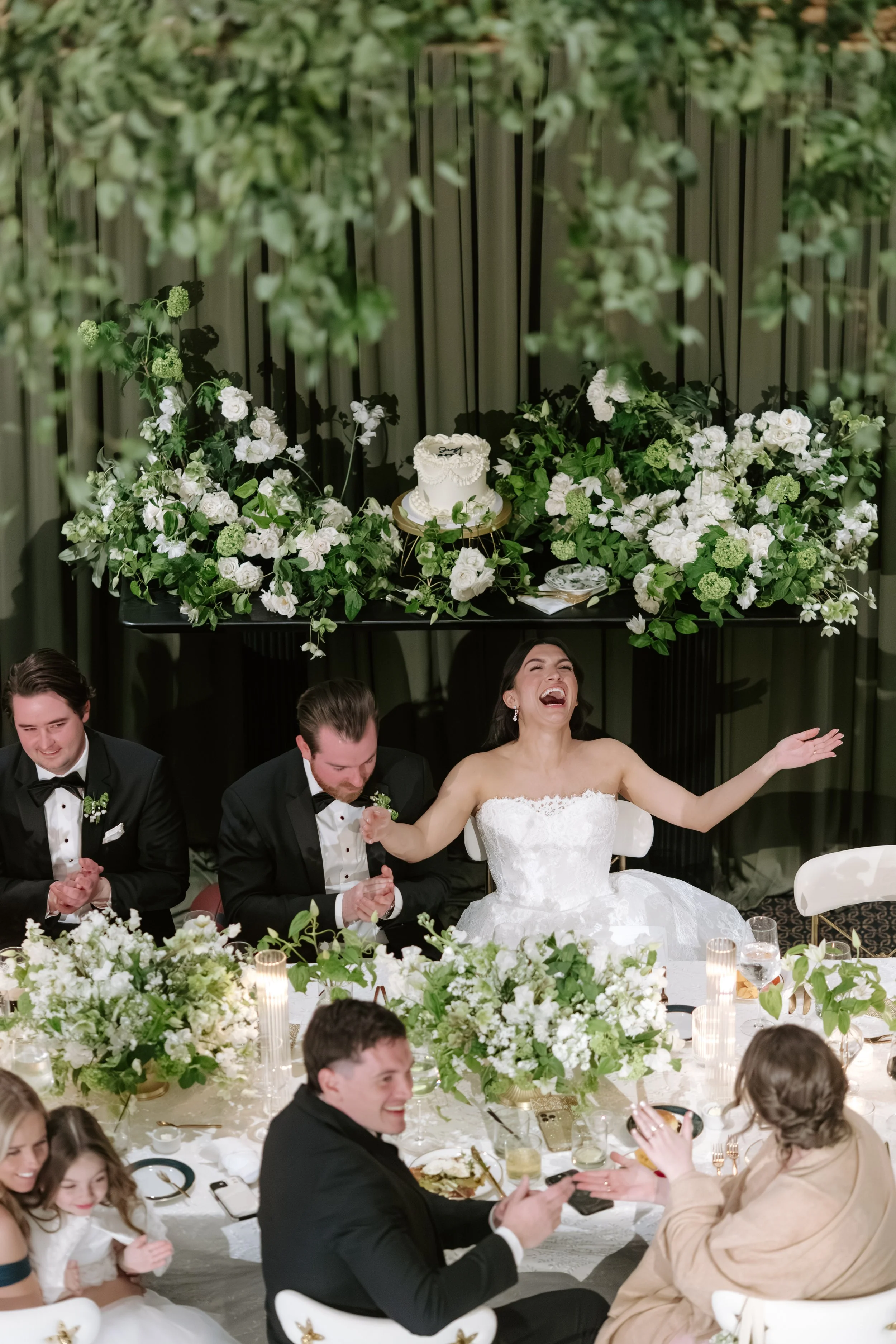 A wedding celebration with smiling bride and groom, surrounded by guests, floral arrangements, and candles on a decorated banquet table.