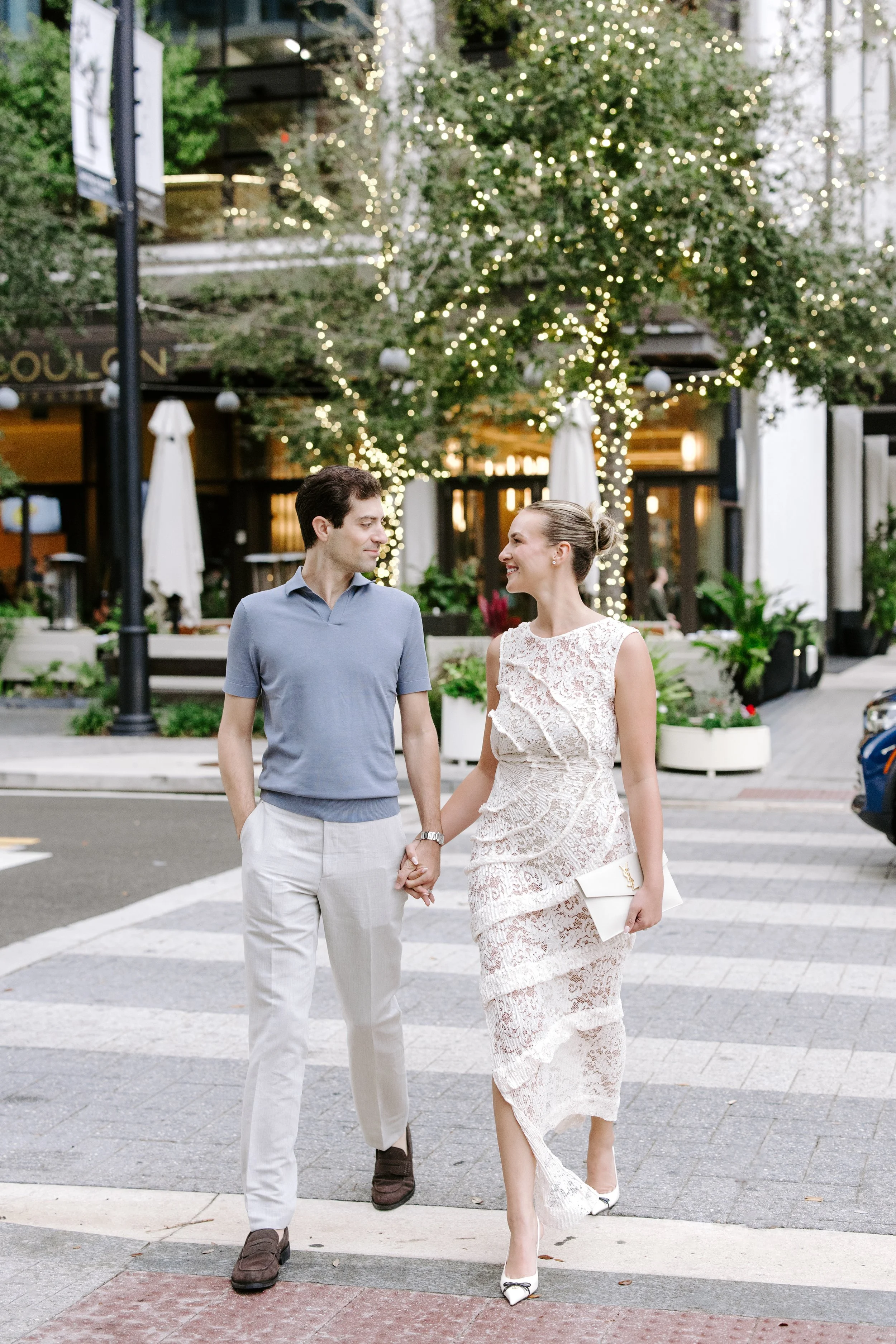A man and woman walking hand in hand on a city sidewalk, the woman wearing a white lace dress and holding a white clutch, the man in a light blue shirt and white trousers, behind a tree decorated with string lights, in an outdoor urban area with umbr