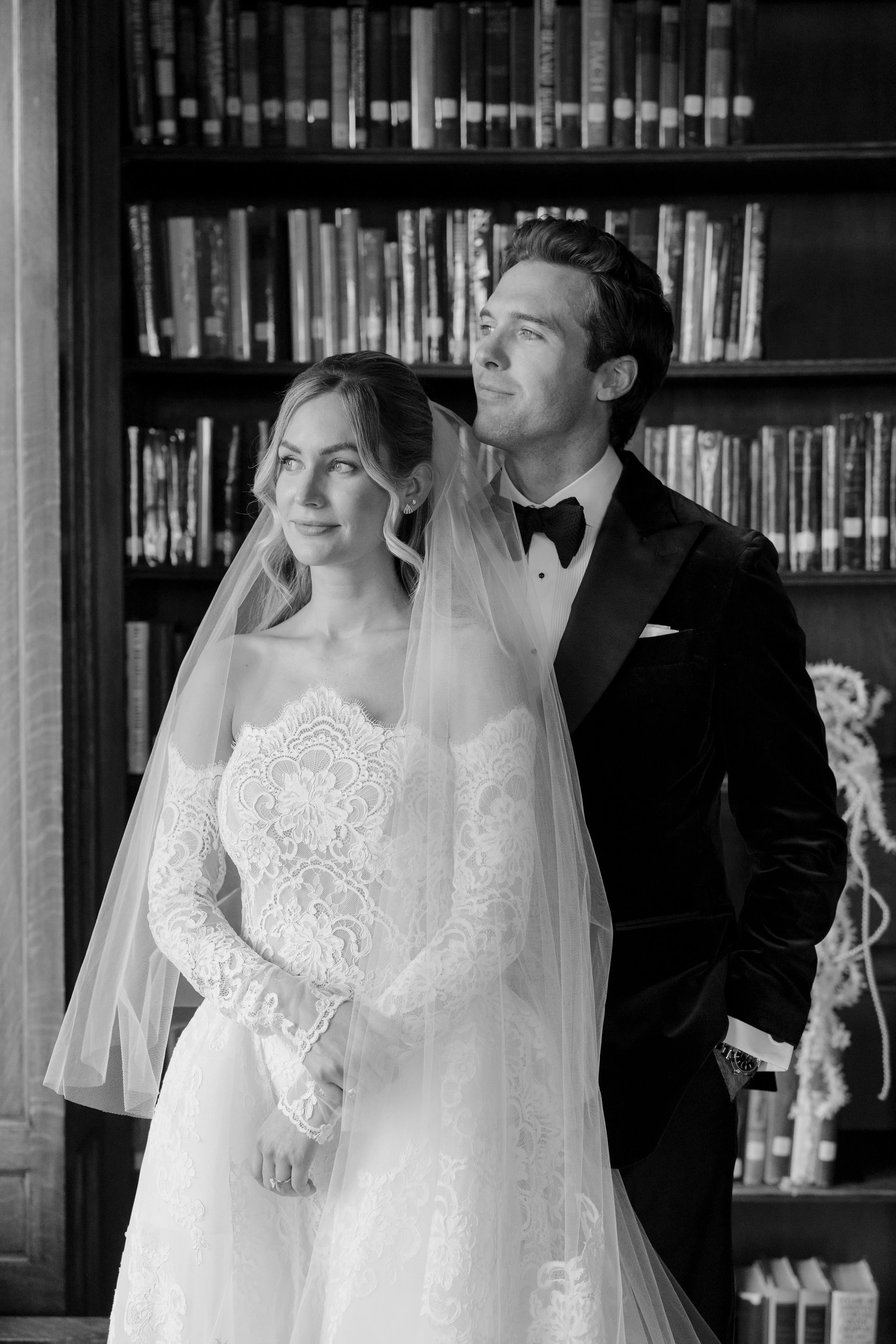 A black and white photo of a bride and groom standing in front of a bookshelf, with the bride wearing a lace wedding gown and veil, and the groom wearing a tuxedo with a bow tie, in a library or study room.