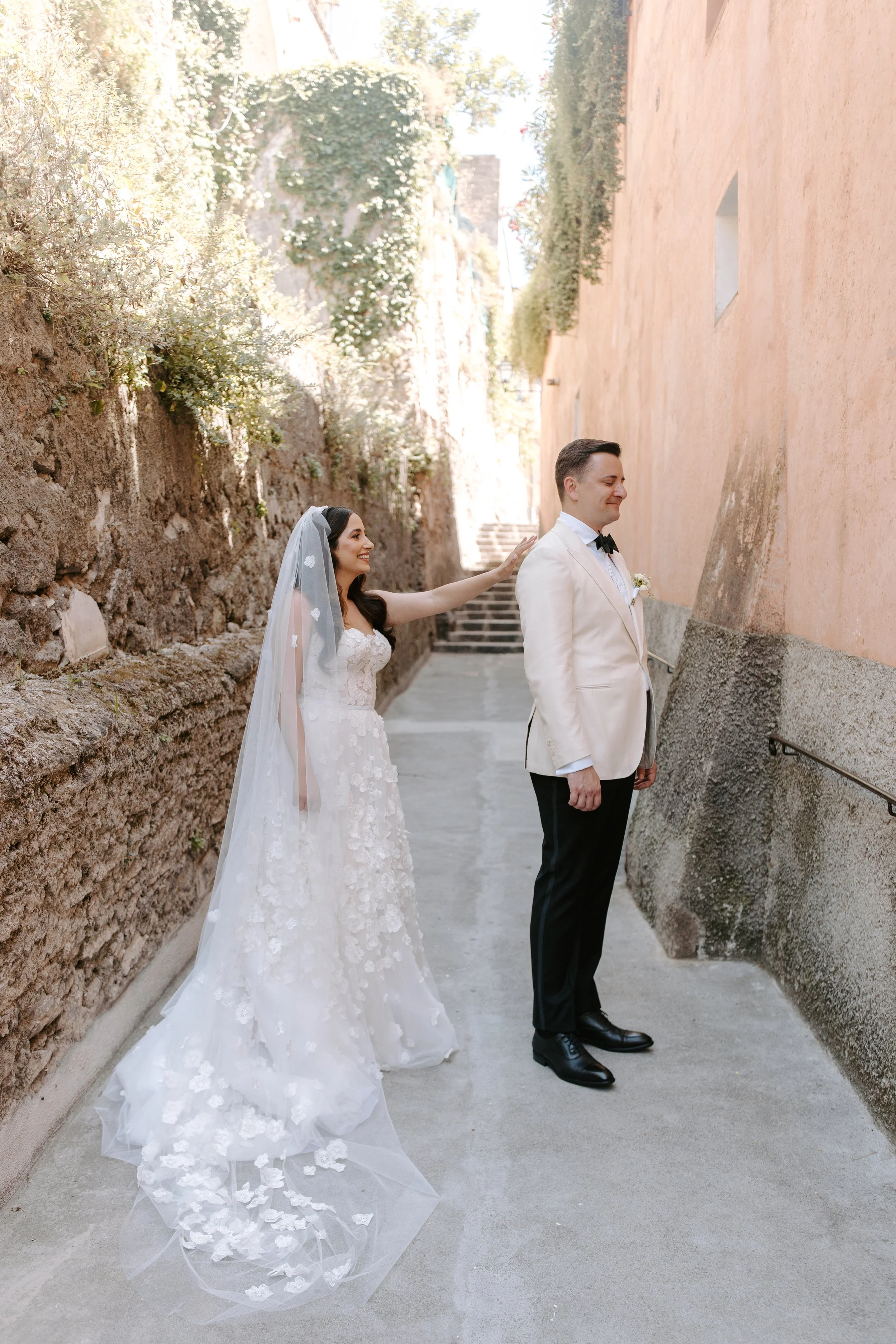 A bride and groom standing in an outdoor alleyway, with the bride touching the groom's shoulder, both smiling, the bride wearing a white wedding dress with a veil and the groom in a cream tuxedo with a black bow tie, stone steps and greenery in the b