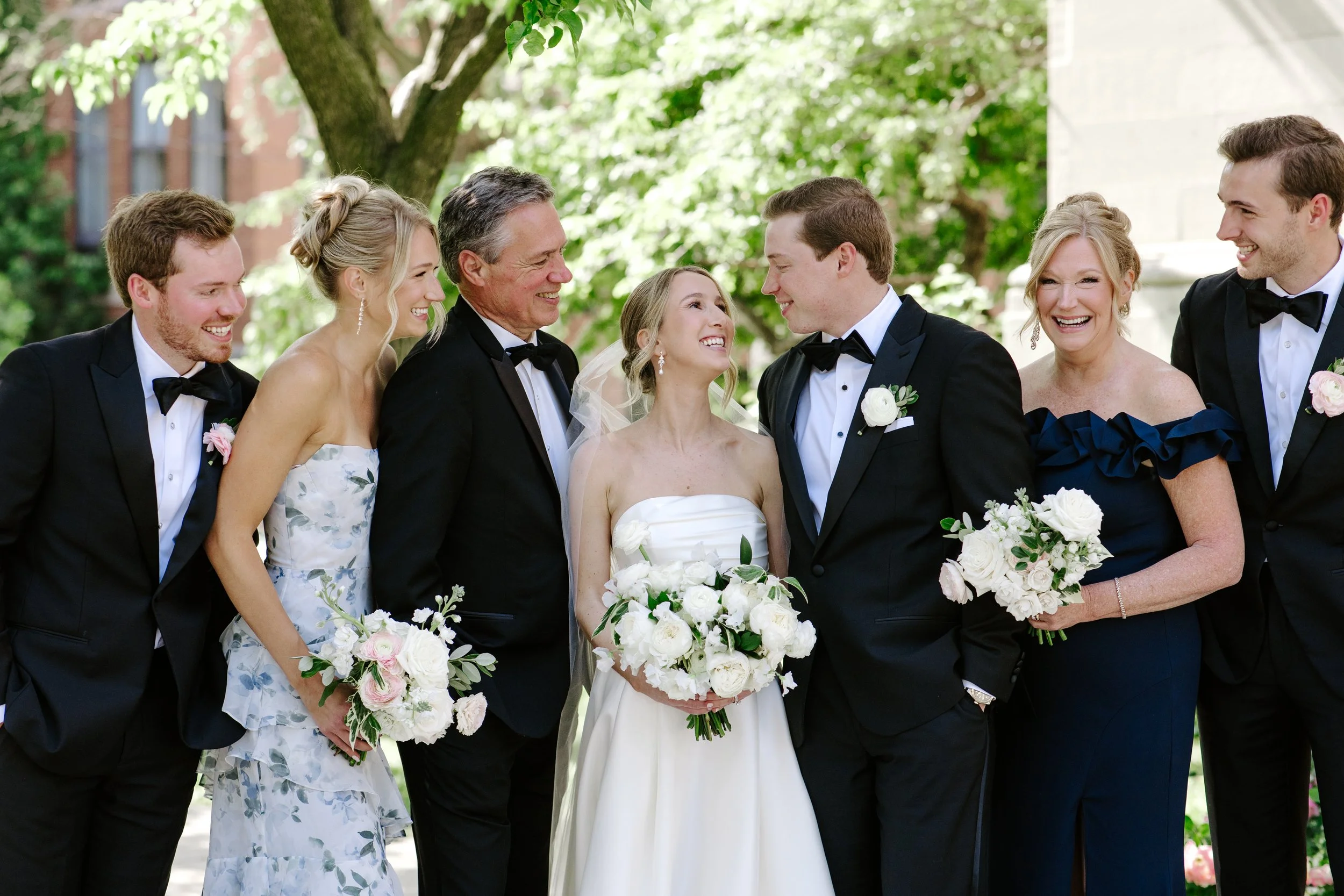 A group of eight people, dressed in formal attire, standing outdoors and smiling at each other during a wedding celebration. The scene takes place under a tree with green leaves, with buildings visible in the background.