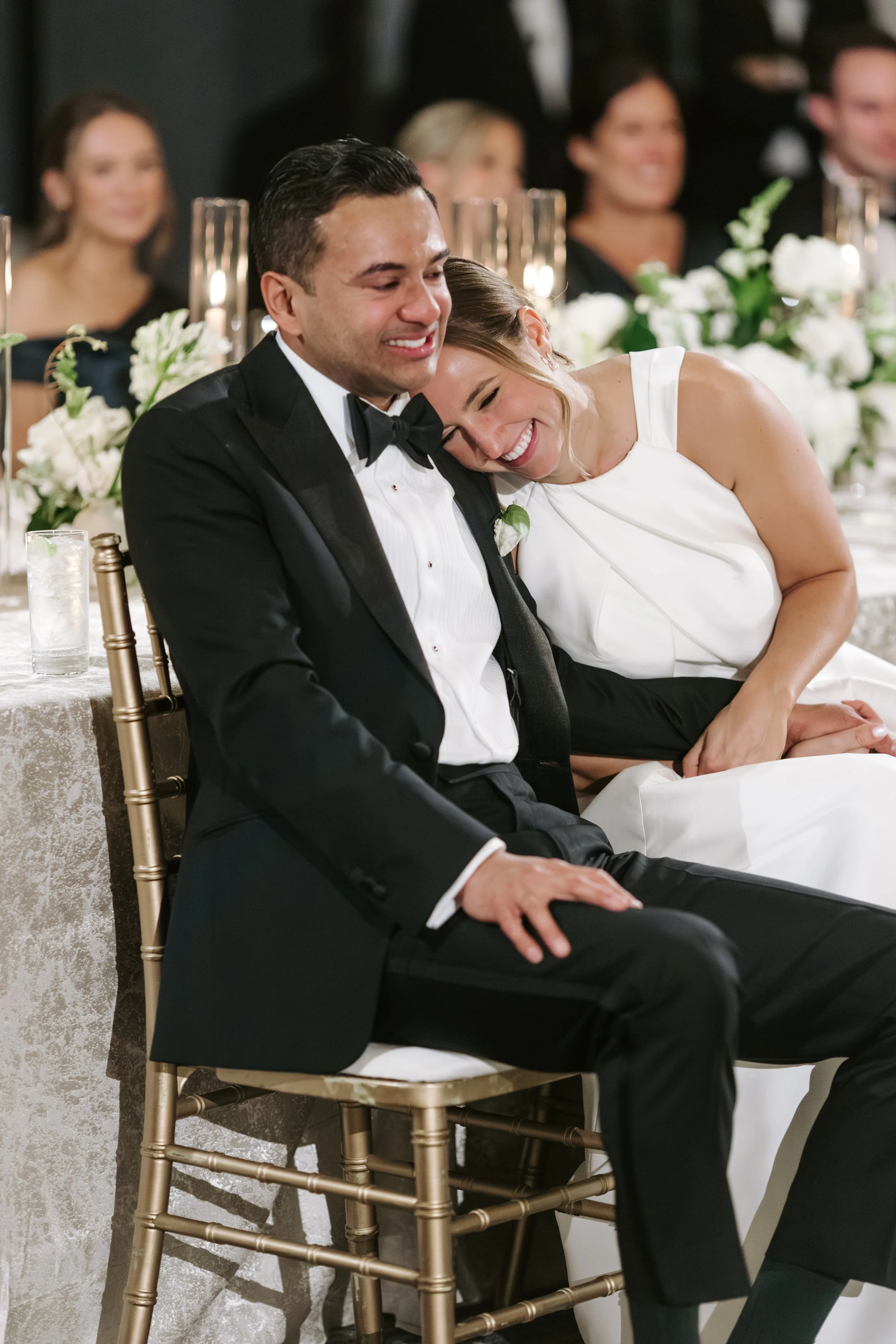 A bride and groom sitting close together at their wedding reception, smiling happily. The bride is resting her head on the groom's shoulder, and they are dressed in formal wedding attire. In the background, guests are seated at a decorated table with