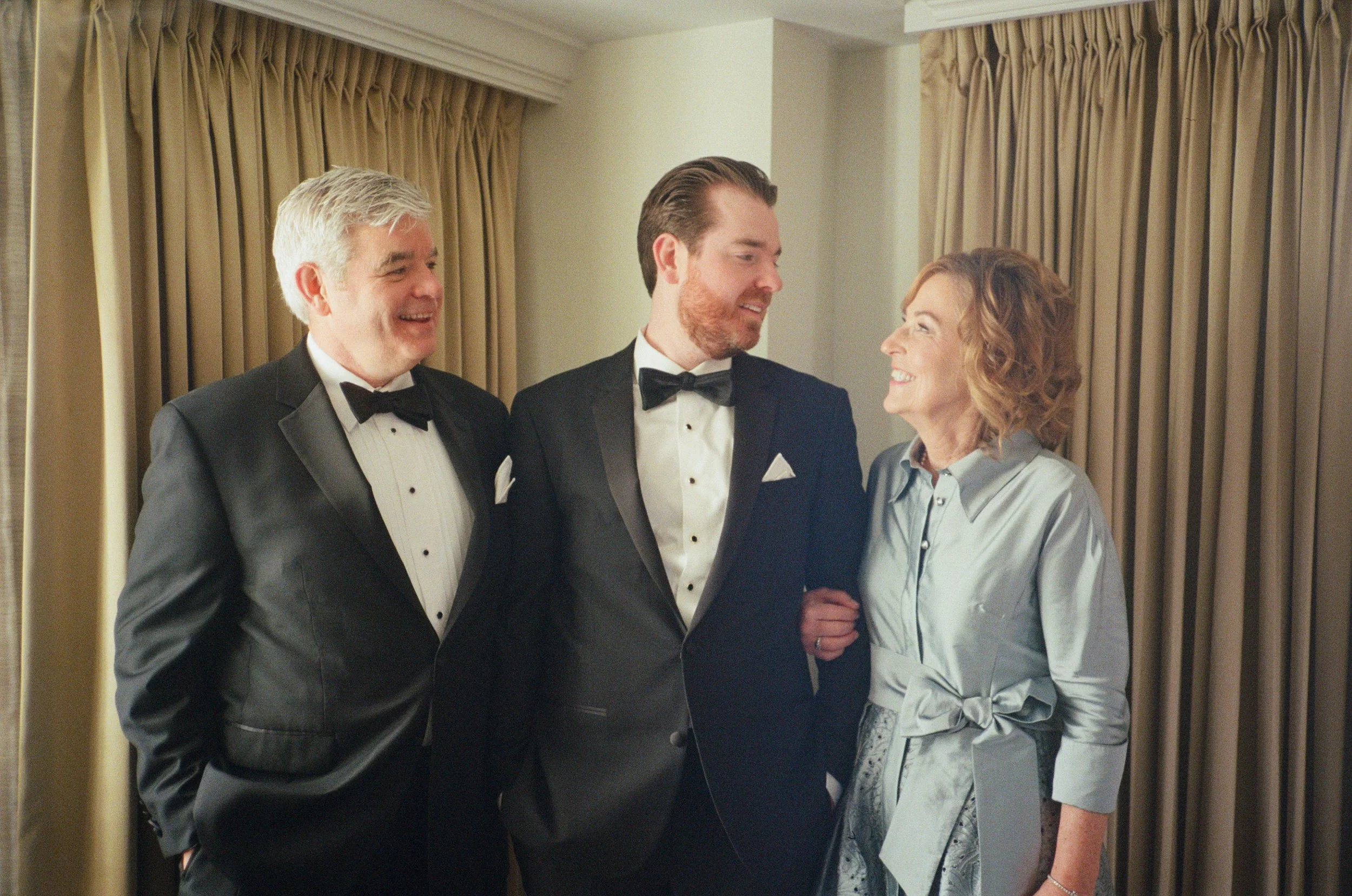 Three people dressed in formal attire, two men in tuxedos and a woman in a satin blouse, standing close together and smiling at each other in front of beige curtains.