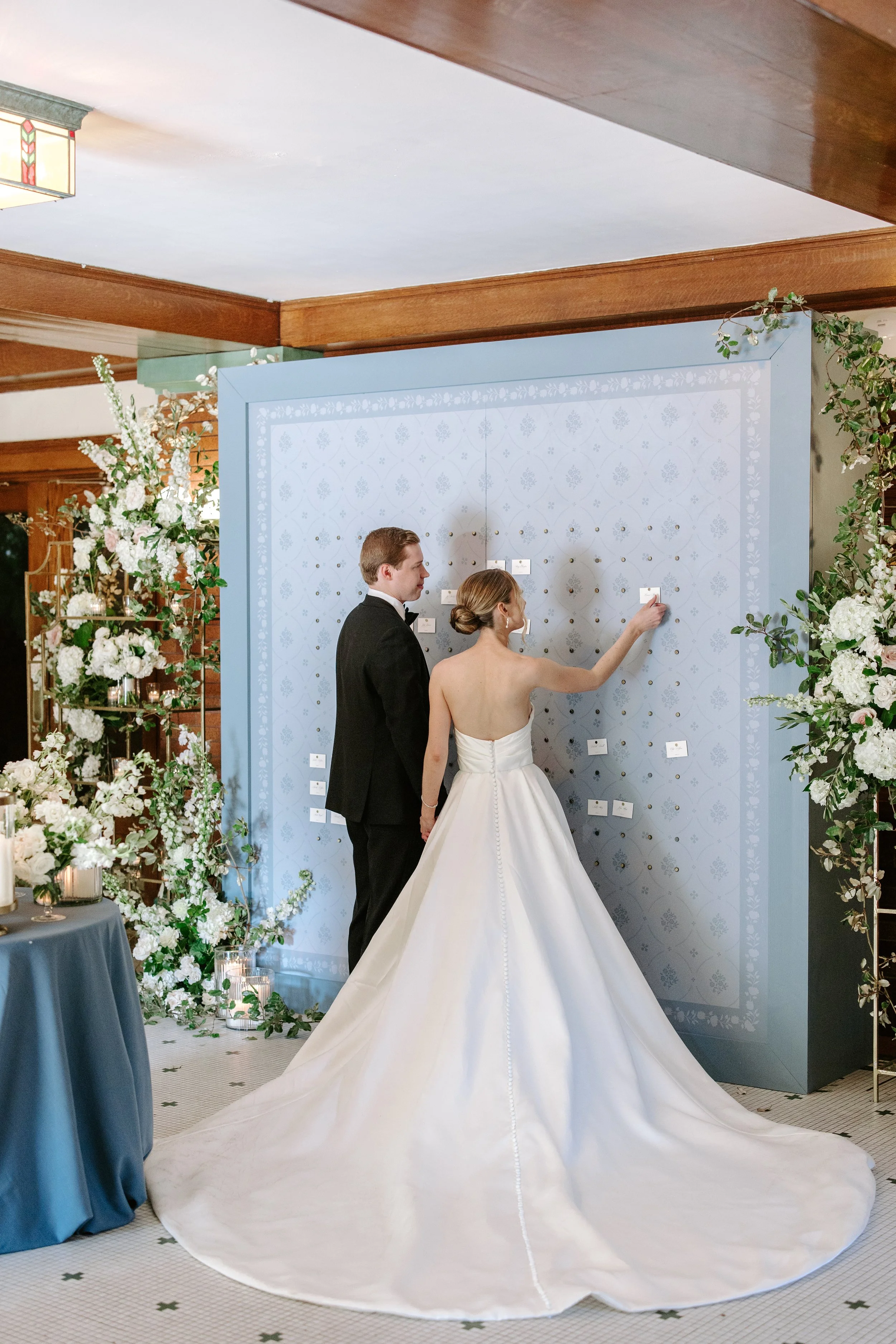 A bride and groom signing a guest book at their wedding, standing in front of a decorated blue wall with floral arrangements on either side.