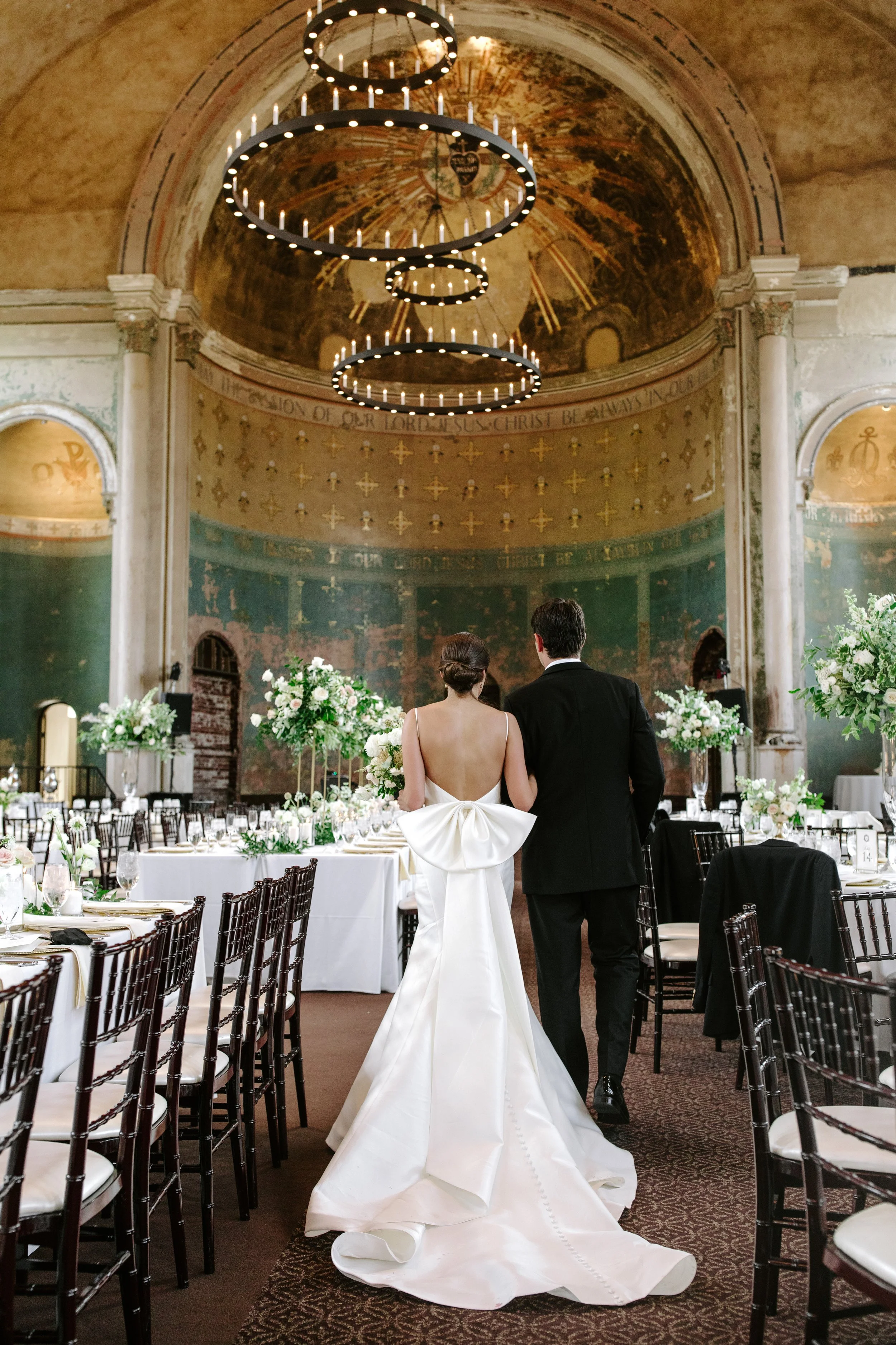 A bride and groom stand together inside a grand, historic building decorated with floral arrangements, preparing for their wedding ceremony. The room features high domed ceilings with ornate paintings, a large circular chandelier, and rows of tables 