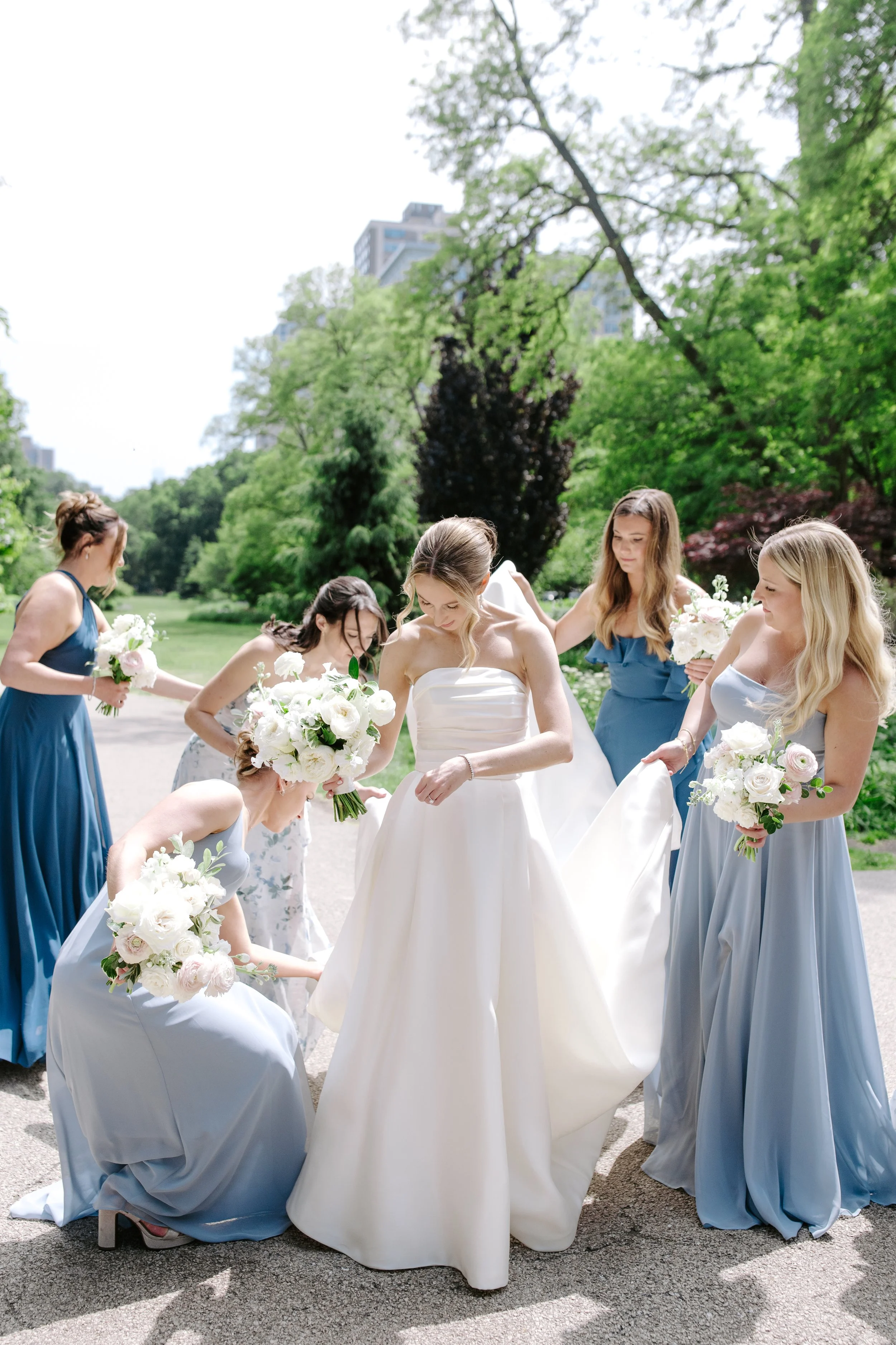 A bride in a white wedding dress surrounded by bridesmaids in blue dresses outdoors on a sunny day with green trees and tall buildings in the background.
