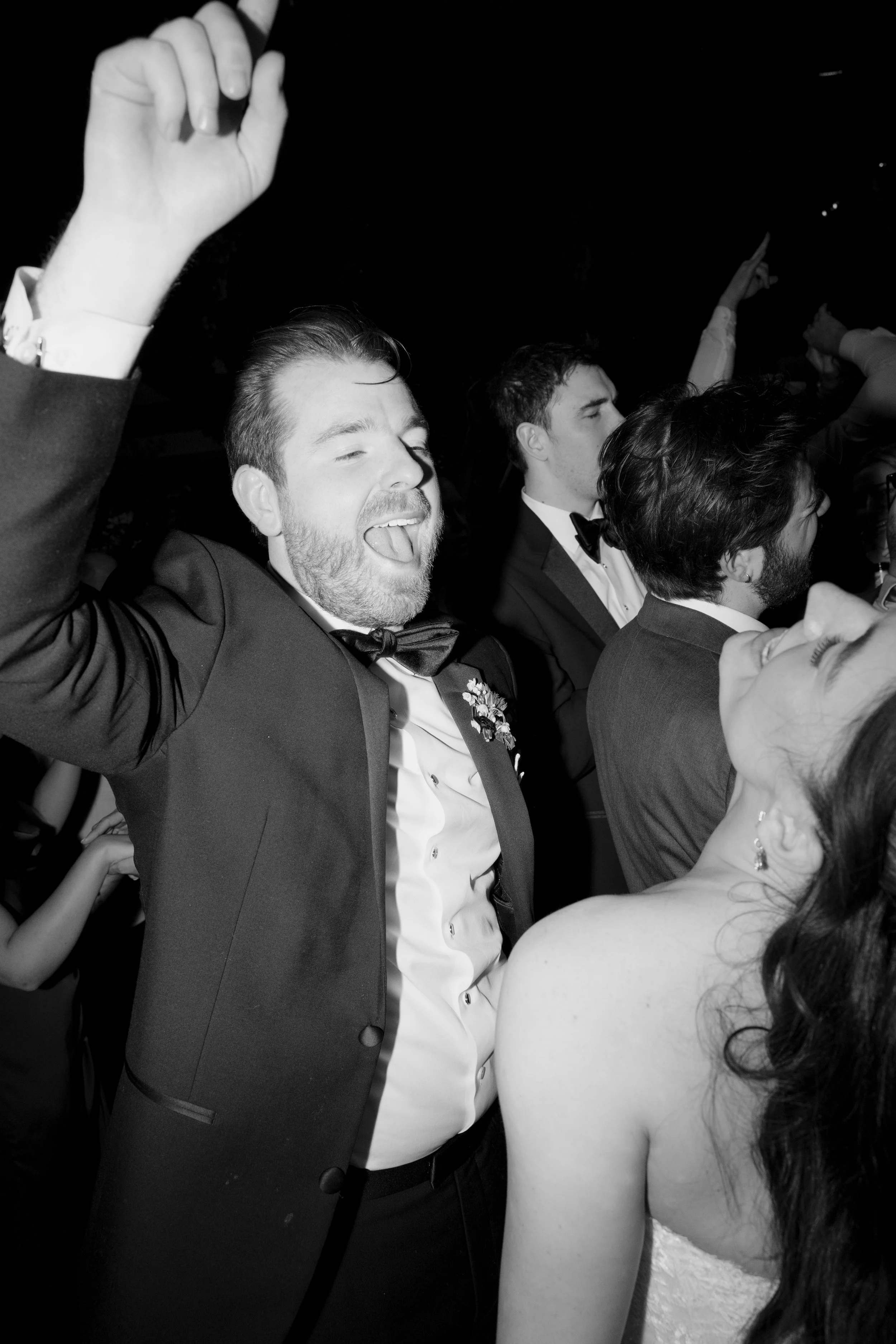 Group of people at a celebration or party, with a man in a tuxedo with a bow tie smiling and raising his hand, surrounded by others in formal attire.