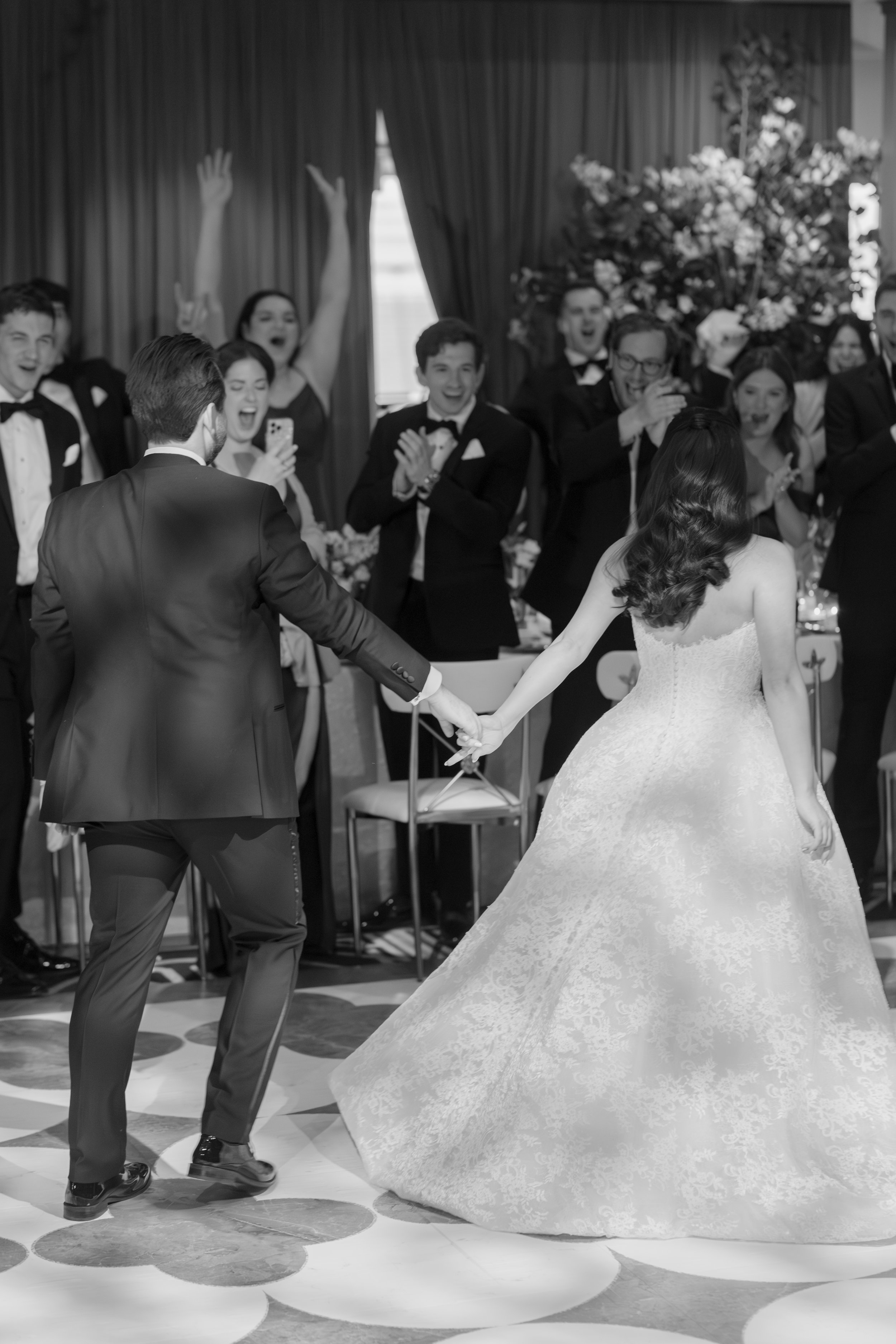 A black and white photo of a wedding reception with a bride and groom dancing, while guests clap and cheer in the background.