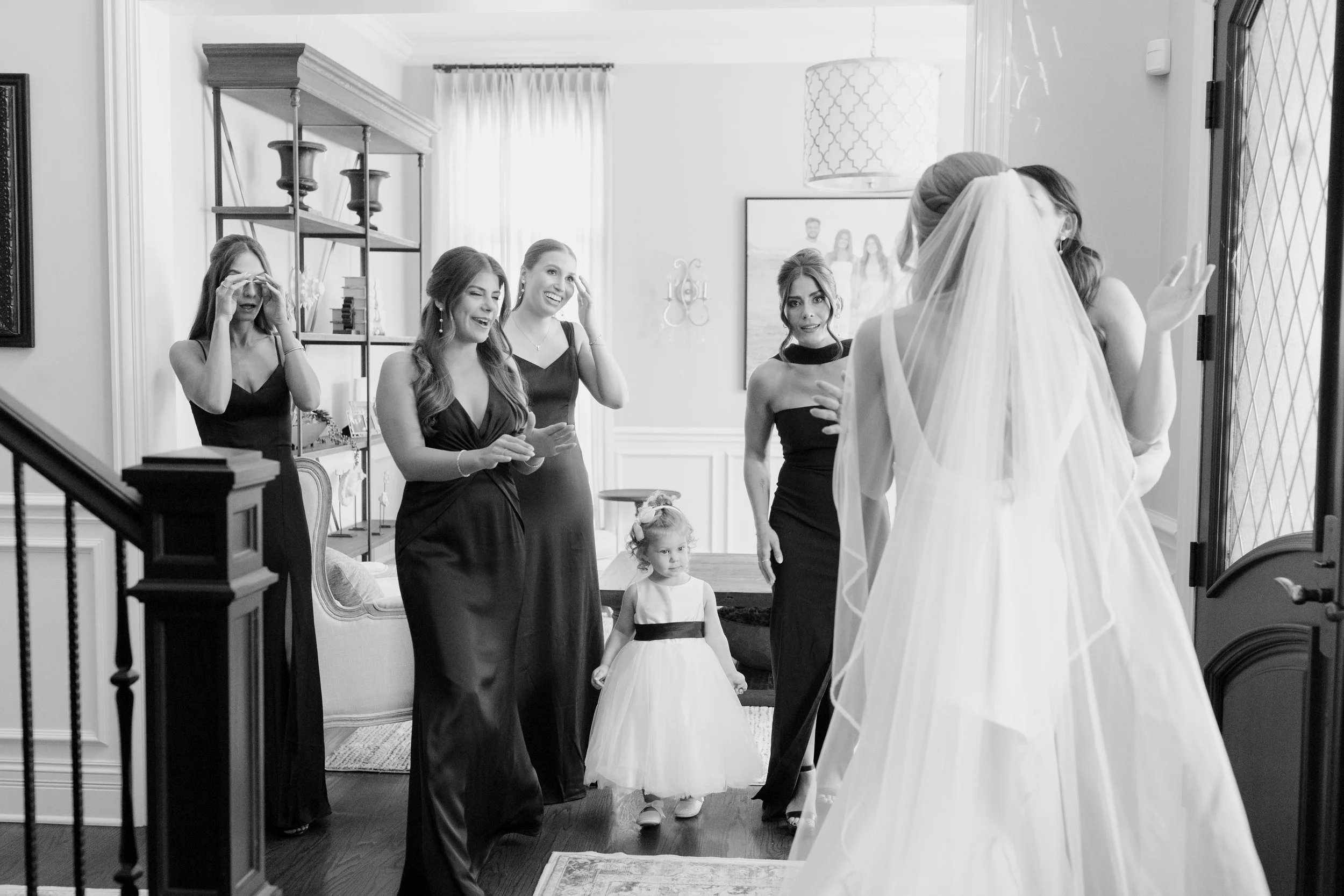 A bride in a wedding dress and veil, facing away, talking with four women in black dresses at a home gathering during the day. Two young girls are also present.