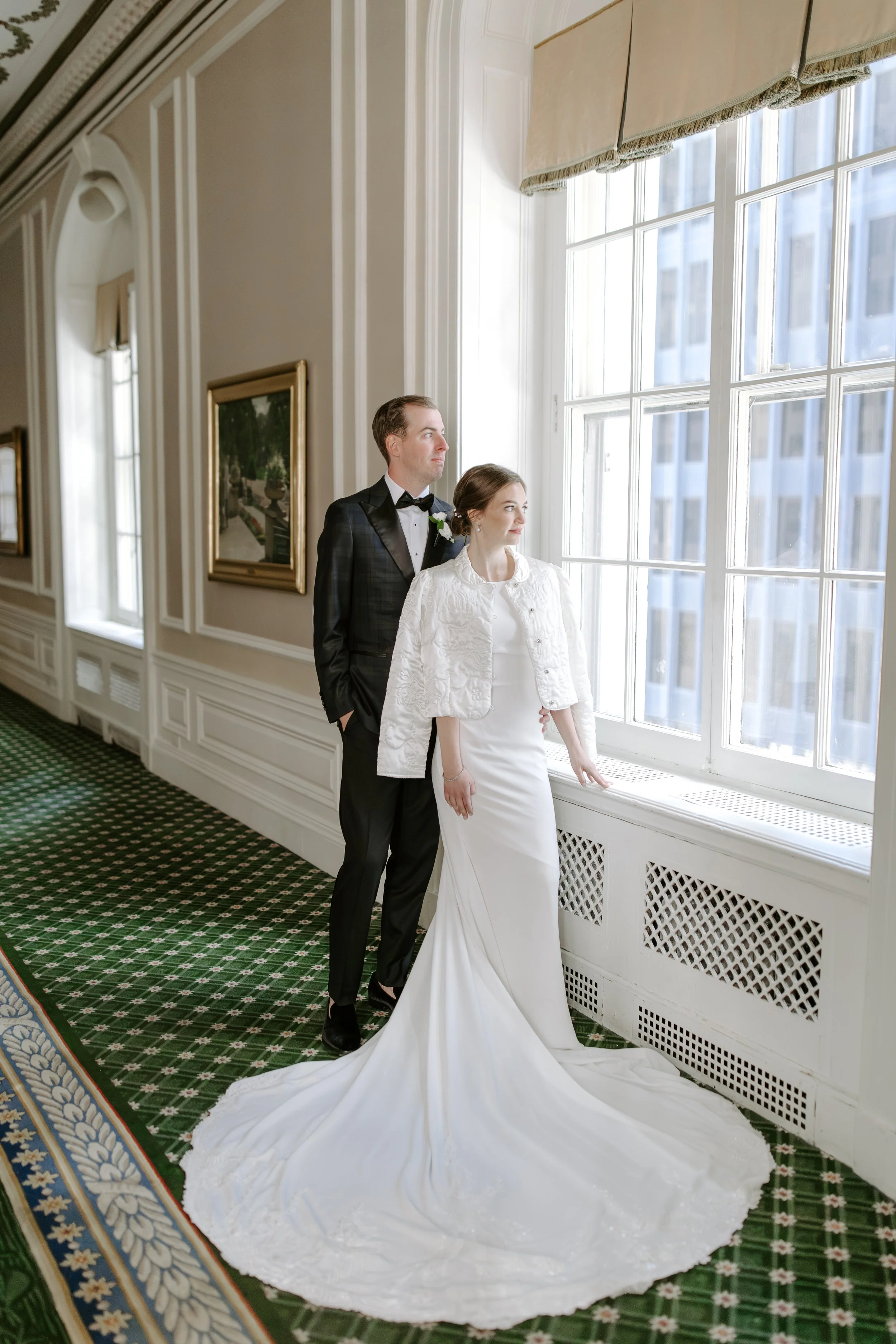 A bride and groom standing indoors near a large window, with the bride wearing a long white wedding dress and the groom in a tuxedo, both gazing out the window.