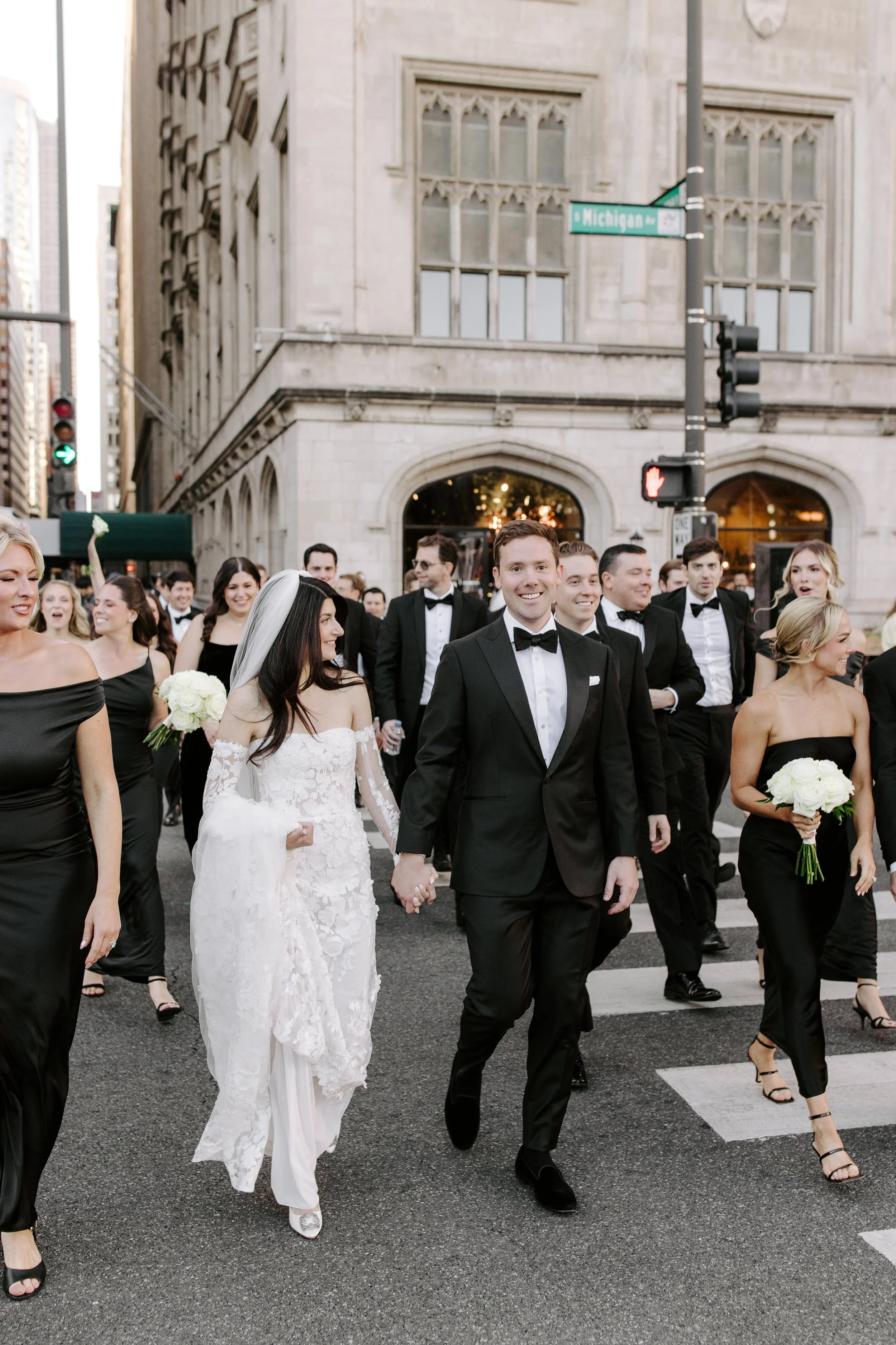 A group of people dressed in formal attire, including a bride in a white wedding dress and a groom in a tuxedo, walking on a city street during a wedding celebration.