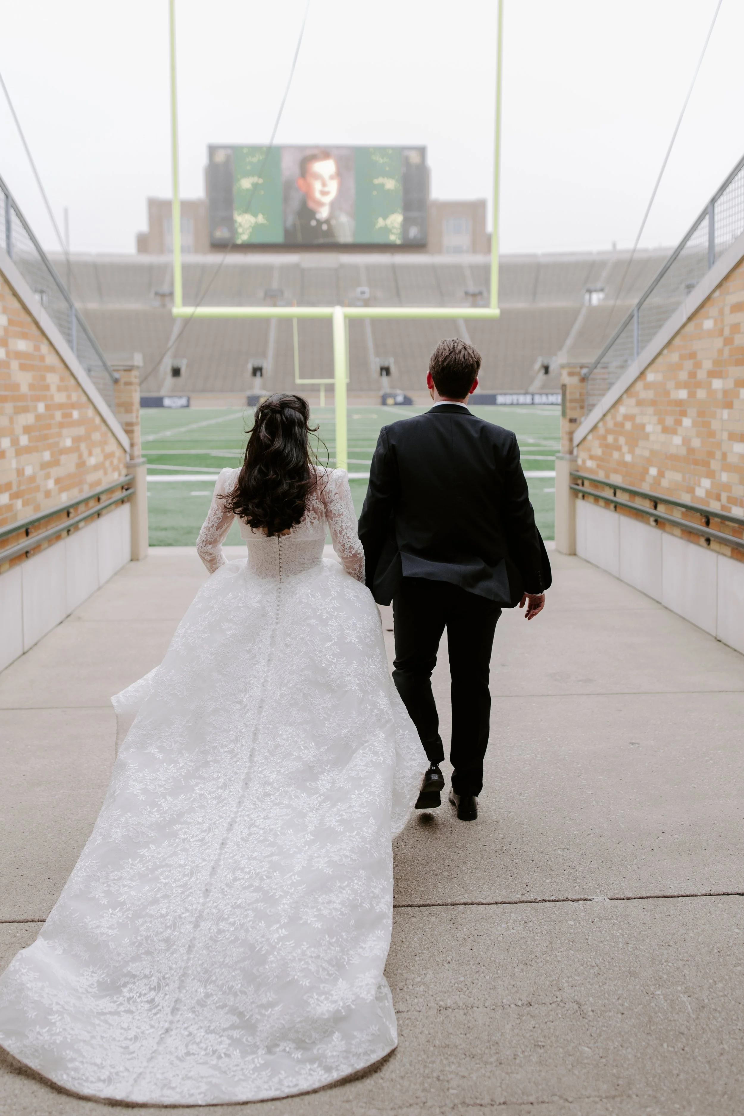 A bride and groom walking through an outdoor stadium tunnel towards the field, with a large screen displaying a portrait of a man in the background.