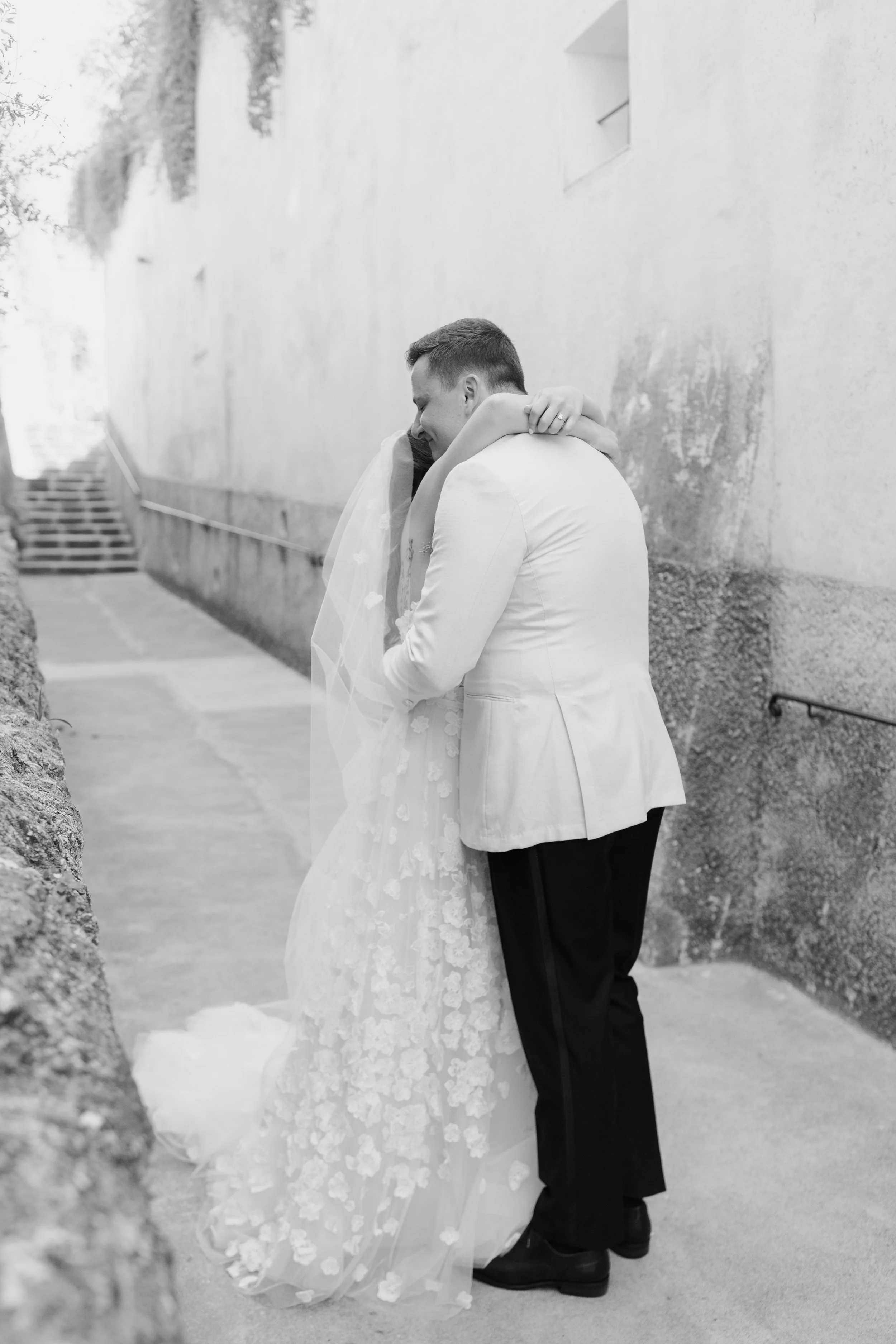 A couple dressed in wedding attire embracing each other outdoors, with the bride’s arm around the groom’s neck and the groom’s hands on the bride’s back, standing on a walkway next to a textured wall.
