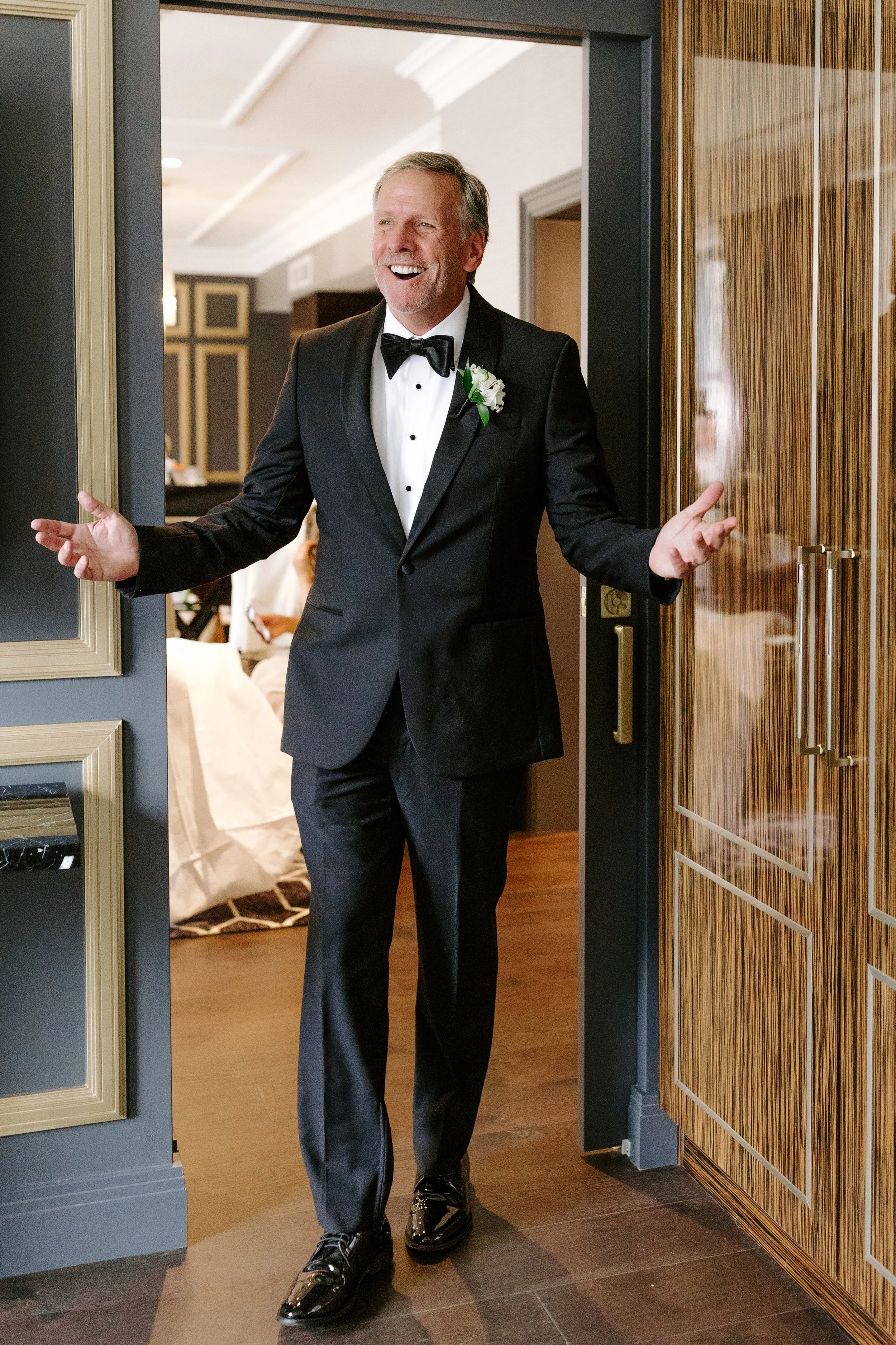 A man in a black tuxedo with a white shirt and black bow tie is smiling and gesturing with his arms as he walks into a room, possibly at a formal event or wedding. The room has wood-paneled walls and warm lighting.