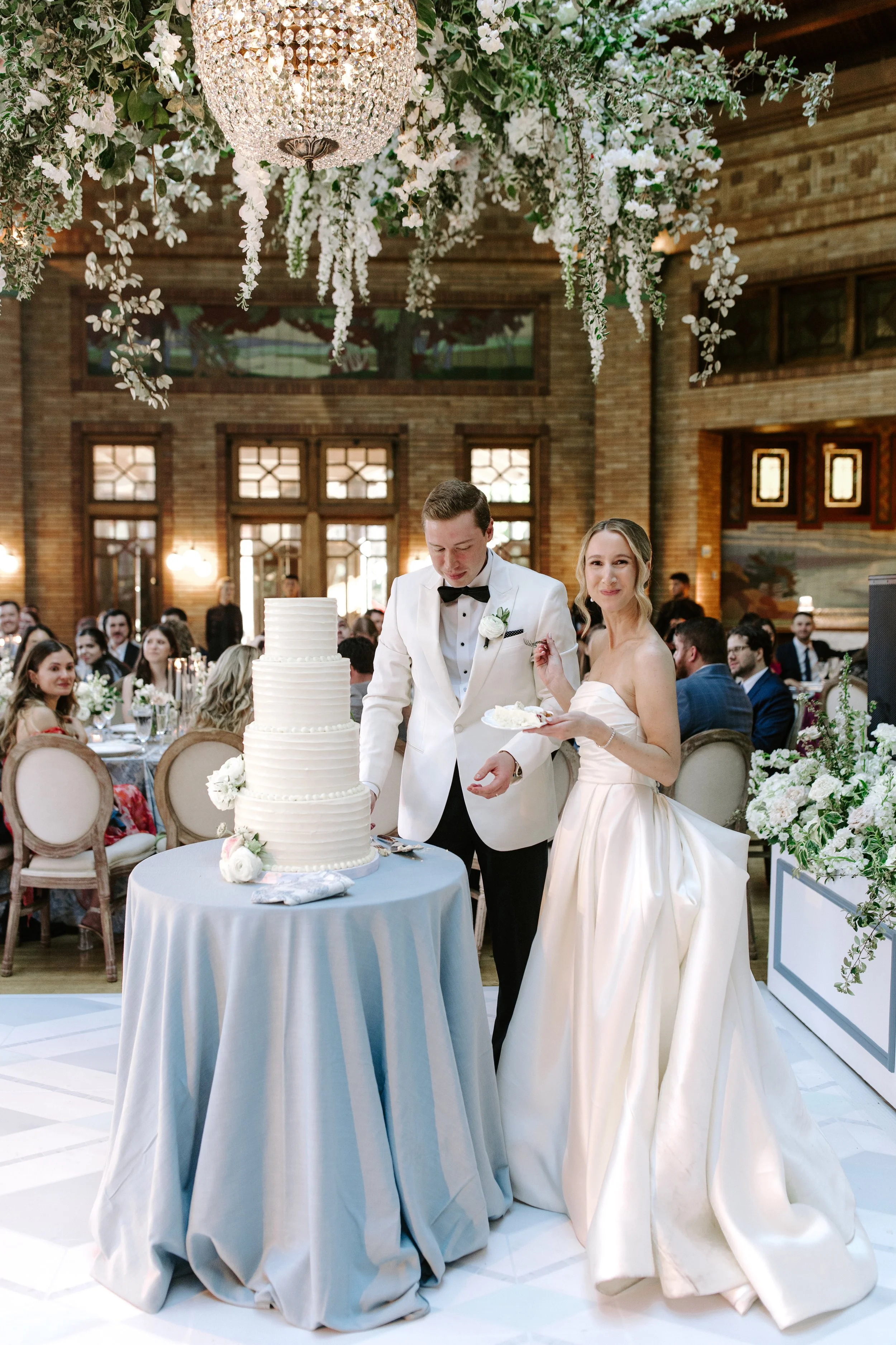 A bride and groom cut a wedding cake at their reception. The bride wears a strapless satin wedding gown, and the groom wears a white tuxedo with a black bow tie. The ceiling is decorated with a large chandelier and hanging white flowers, and guests are seated at decorated tables.