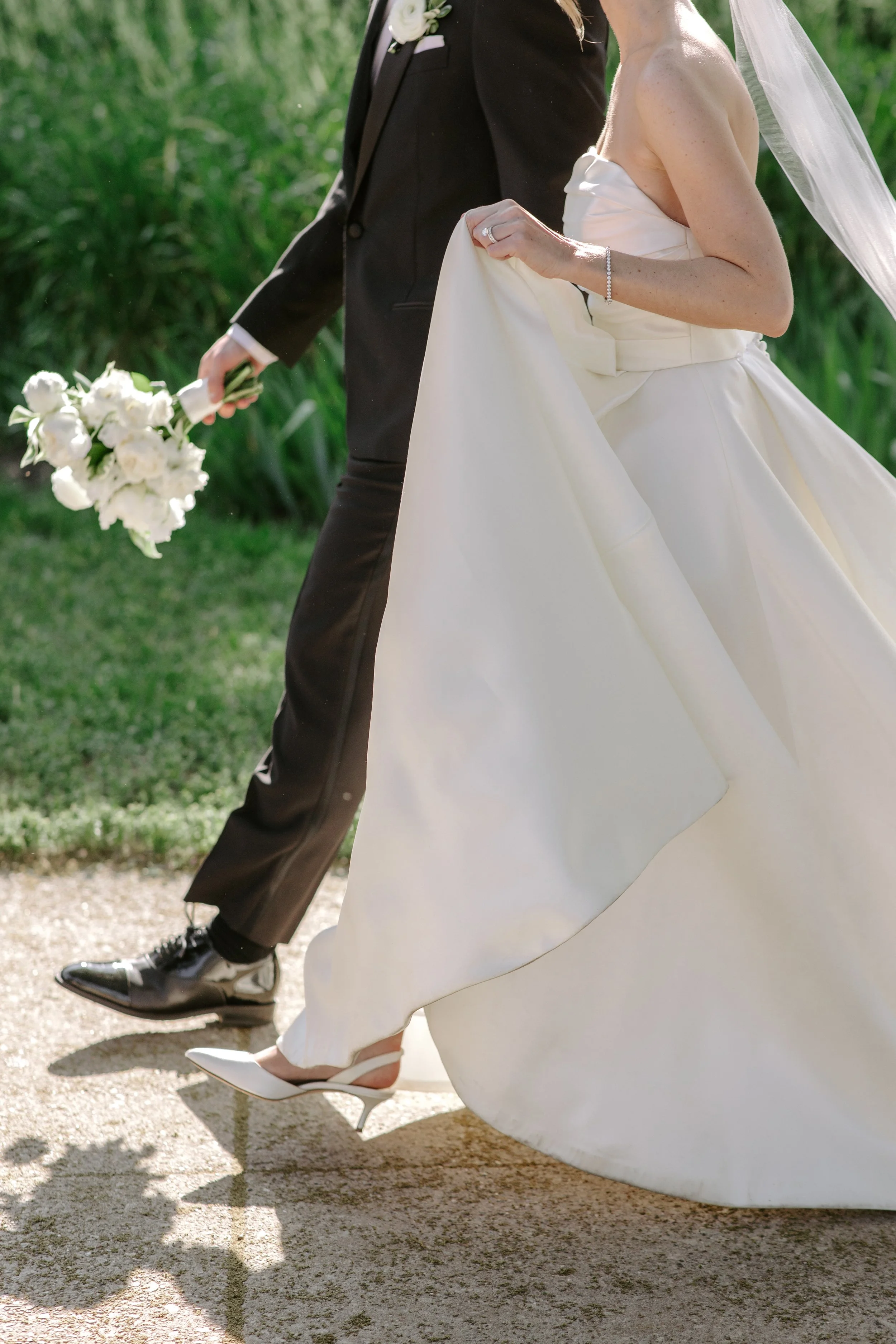 A bride and groom walking outdoors, holding hands. The bride is in a white wedding dress and heels, and the groom is in a black suit and dress shoes. The groom is holding a bouquet of white flowers.