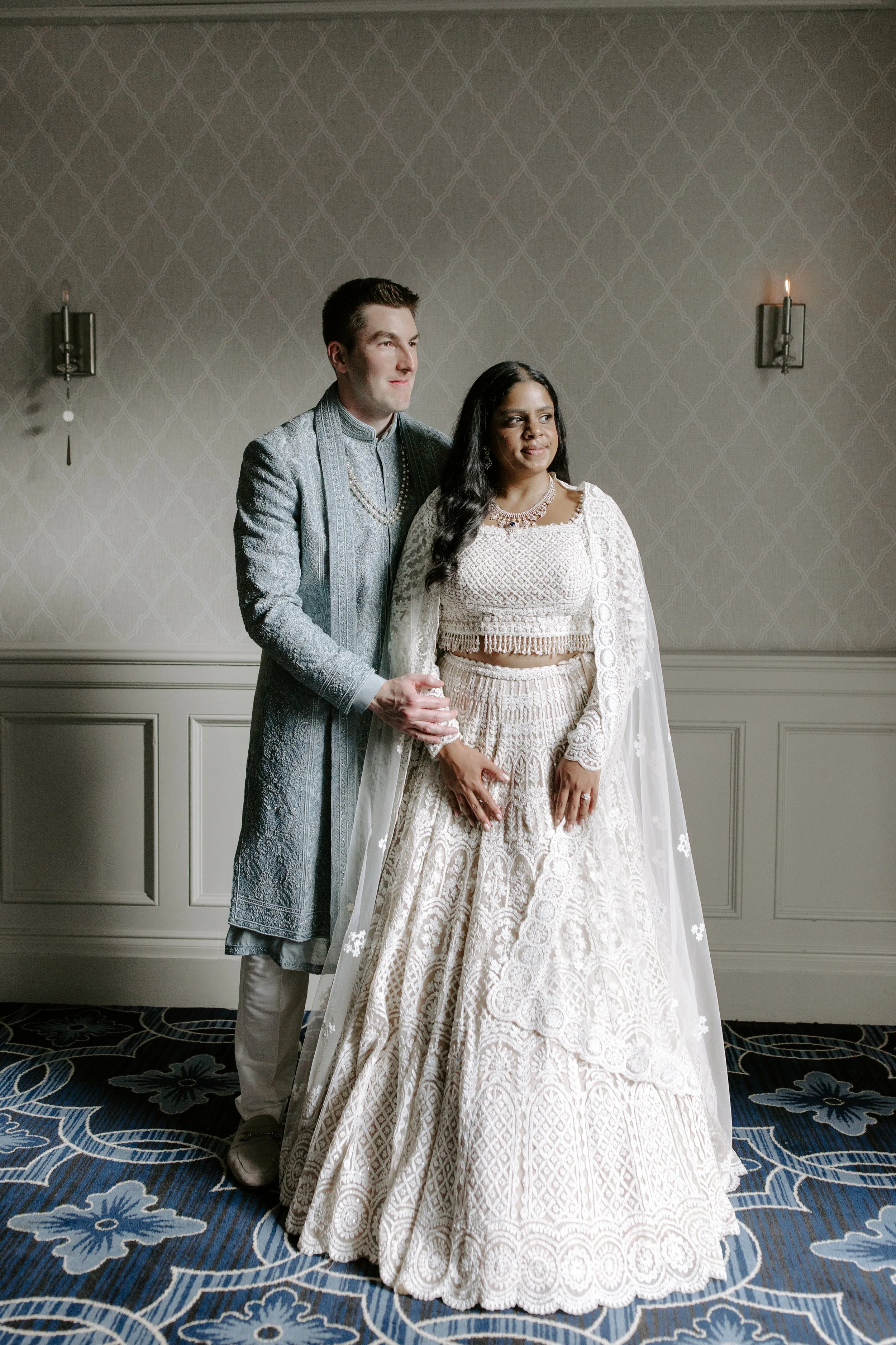 A wedding couple standing in a decorated room, the man in a silver traditional outfit with a pearl necklace, and the woman in a white lace wedding gown with jewelry and a shawl. They are holding hands and looking to the right.