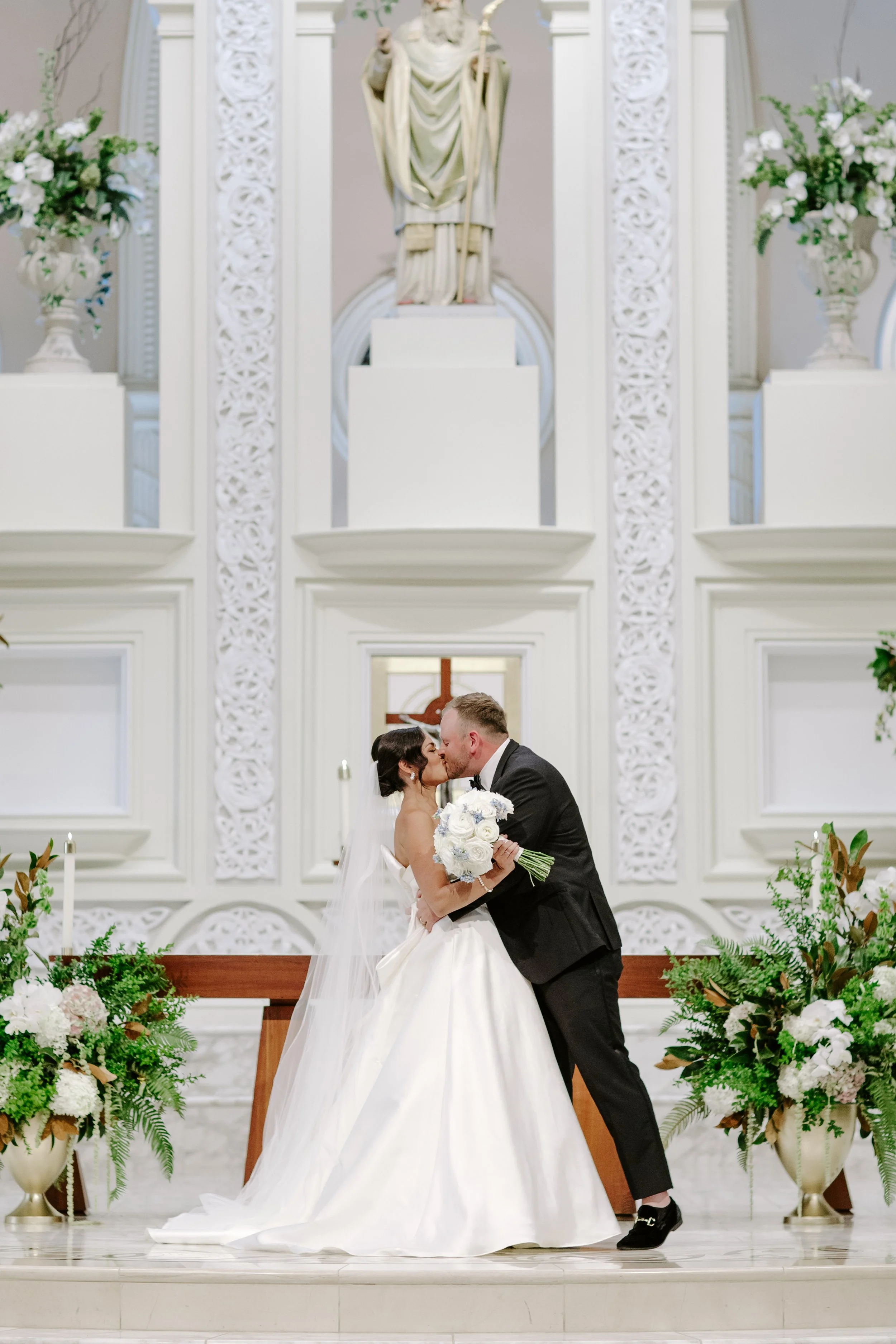 A newlywed couple kissing at the altar inside a church. The bride is wearing a white wedding gown and holding a bouquet of white flowers. The groom is dressed in a black tuxedo. The background features white decorative architecture and floral arrange
