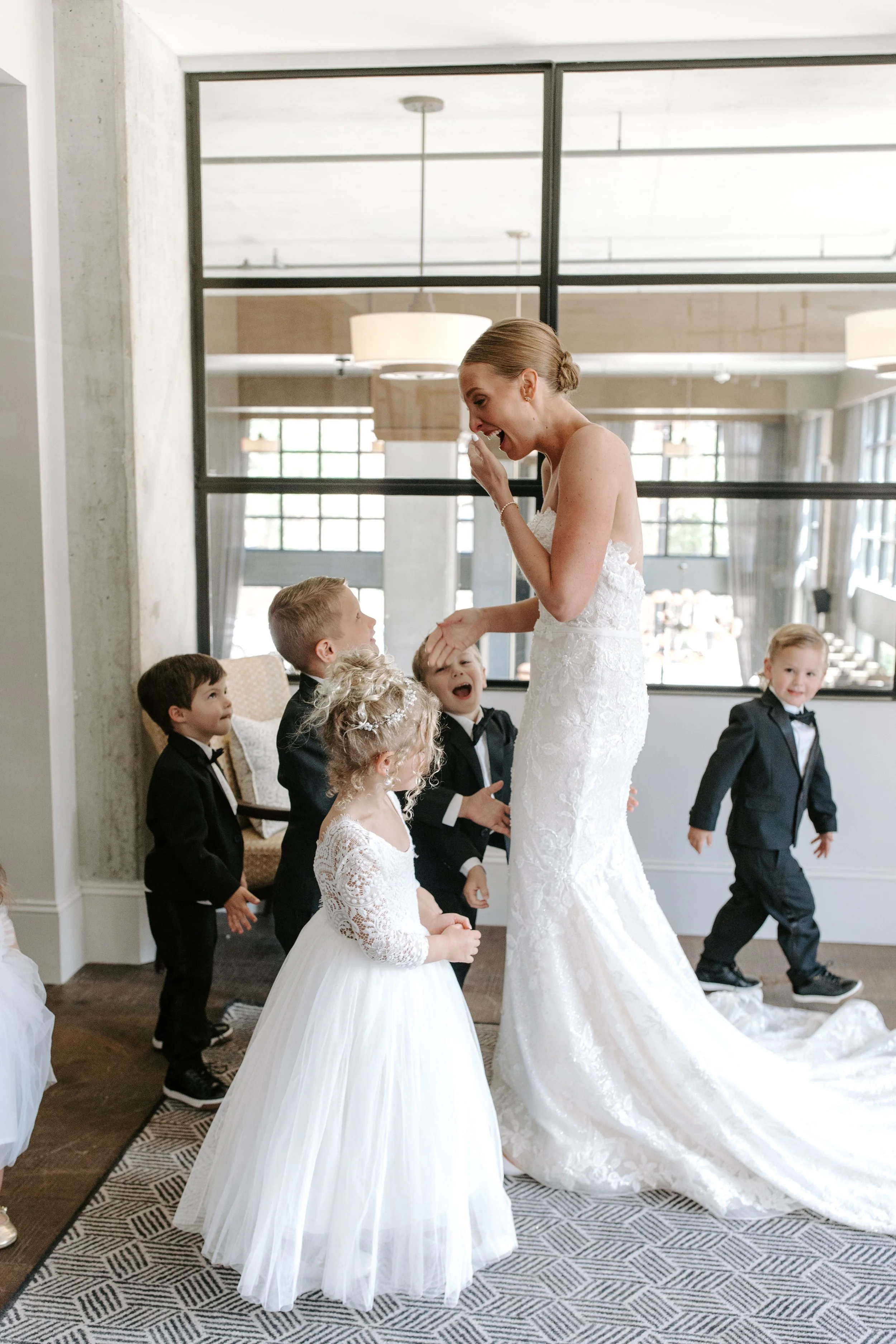 A woman dressed in a white wedding gown, smiling and covering her mouth with her hand, interacting with young children in formal attire in a bright, modern indoor space with large windows and hanging lights.