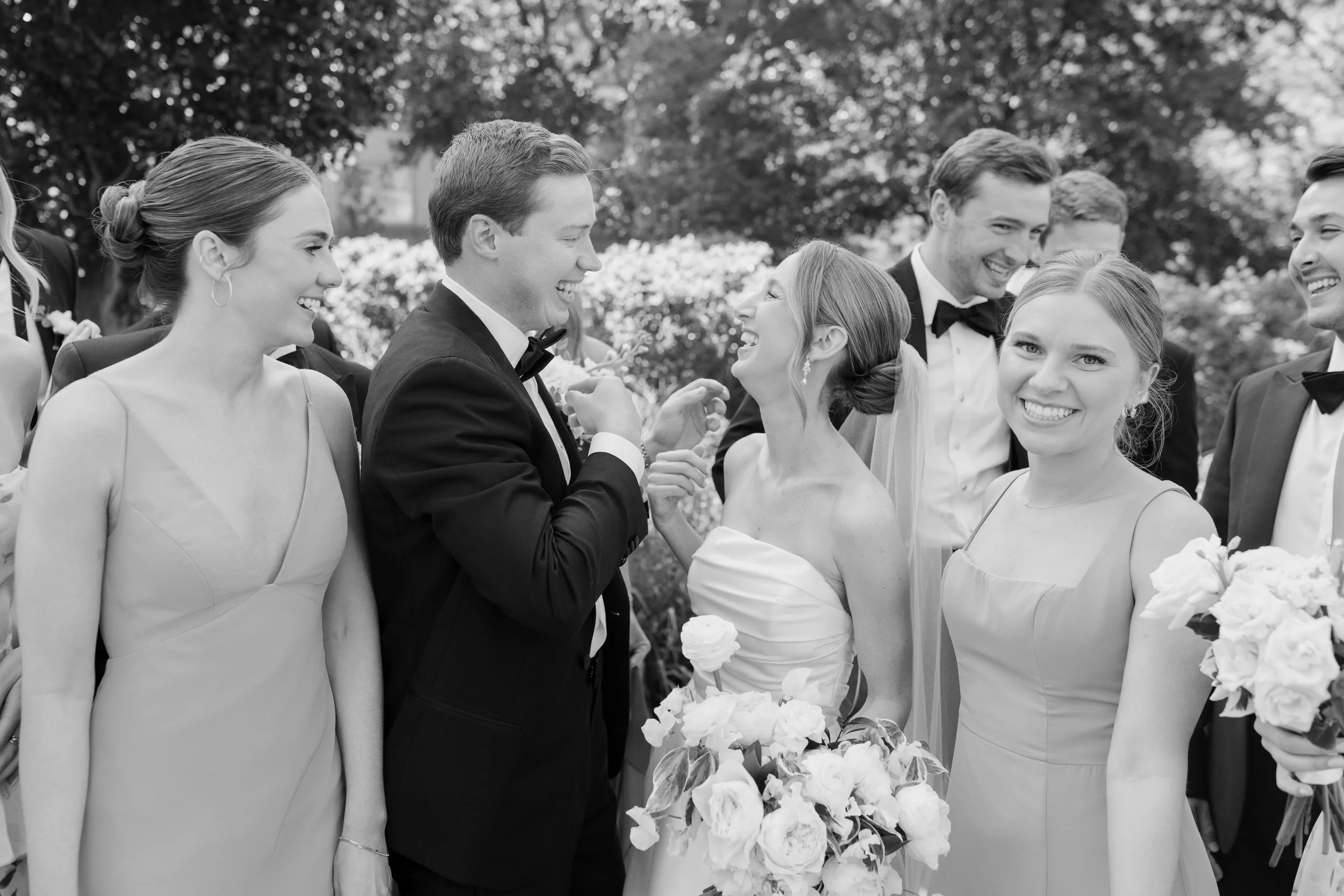 A black and white photograph of a group of wedding guests smiling and enjoying the celebration outdoors. The bride and groom are at the center, holding hands and looking at each other happily, surrounded by friends and family.