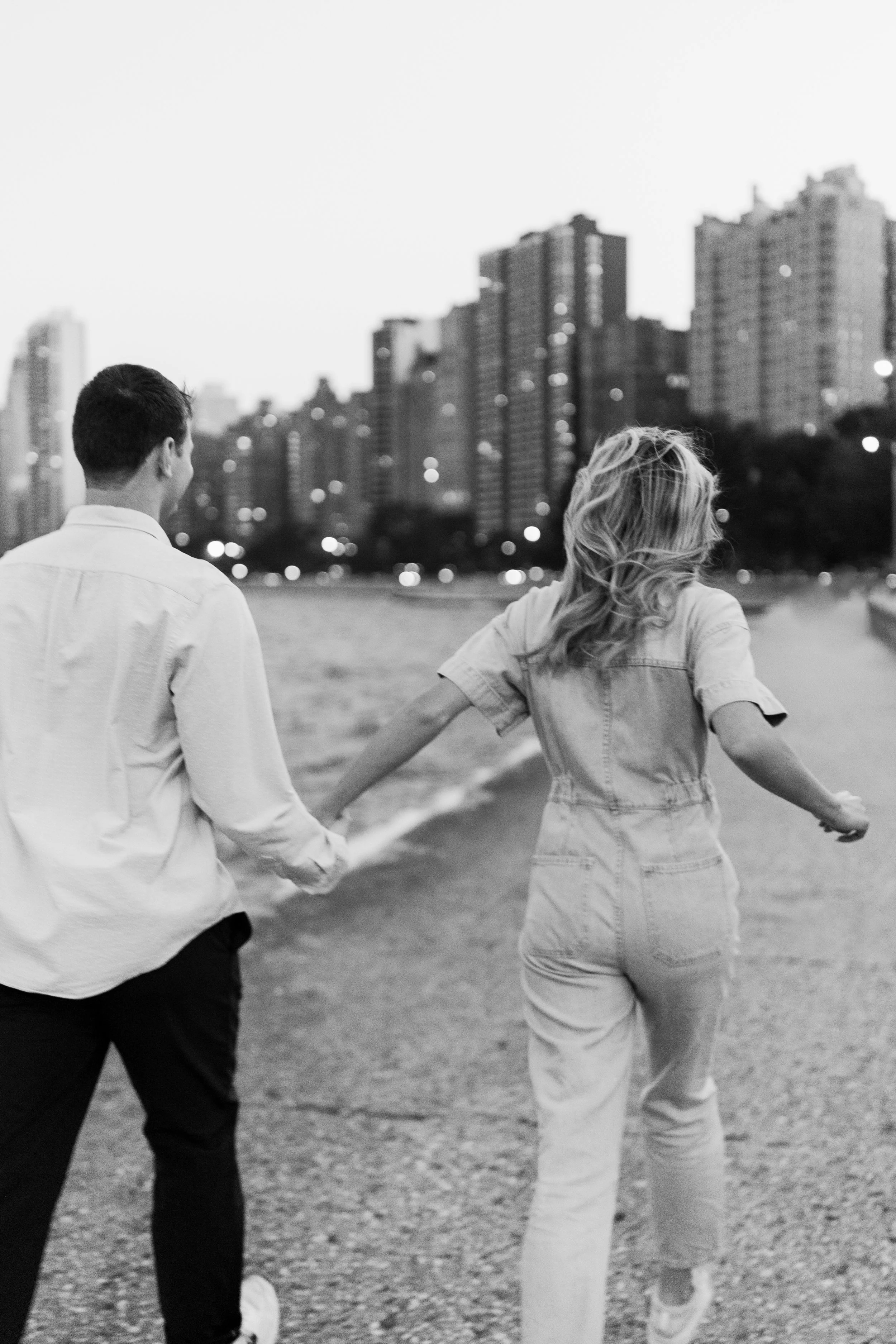 A black and white photo of a young couple holding hands and walking outdoors near city high-rise buildings, with the woman on the right and the man on the left.