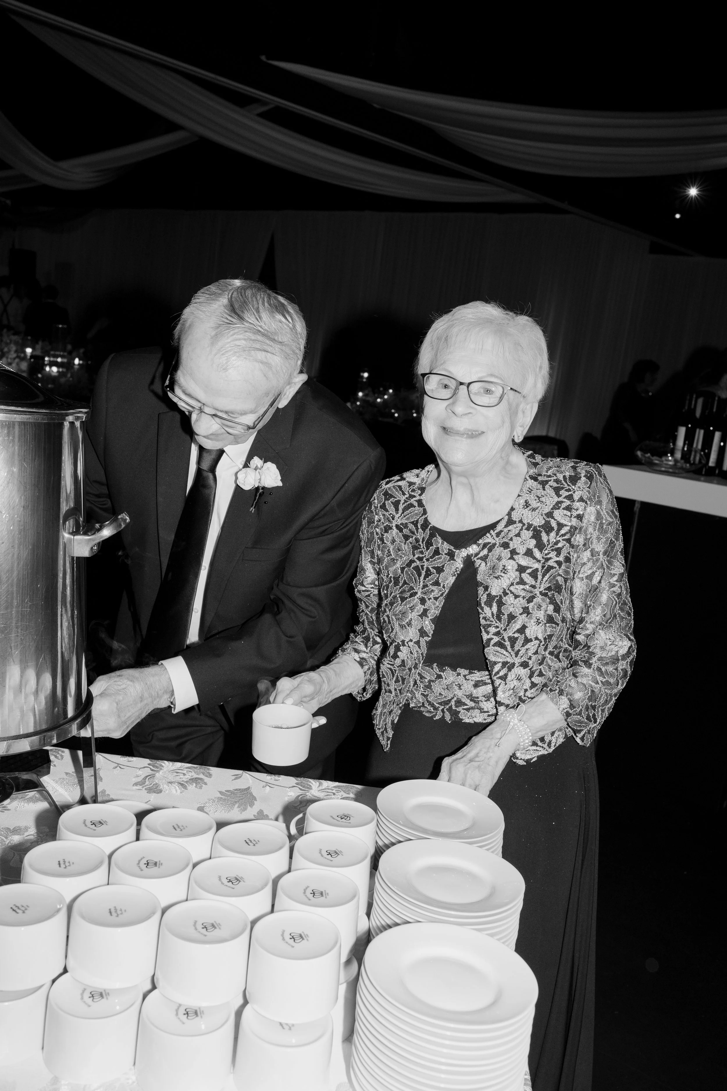 An elderly woman in a patterned jacket and glasses, smiling, standing next to an elderly man in a suit as they serve beverages at a buffet table with cups and plates, in a decorated event space.