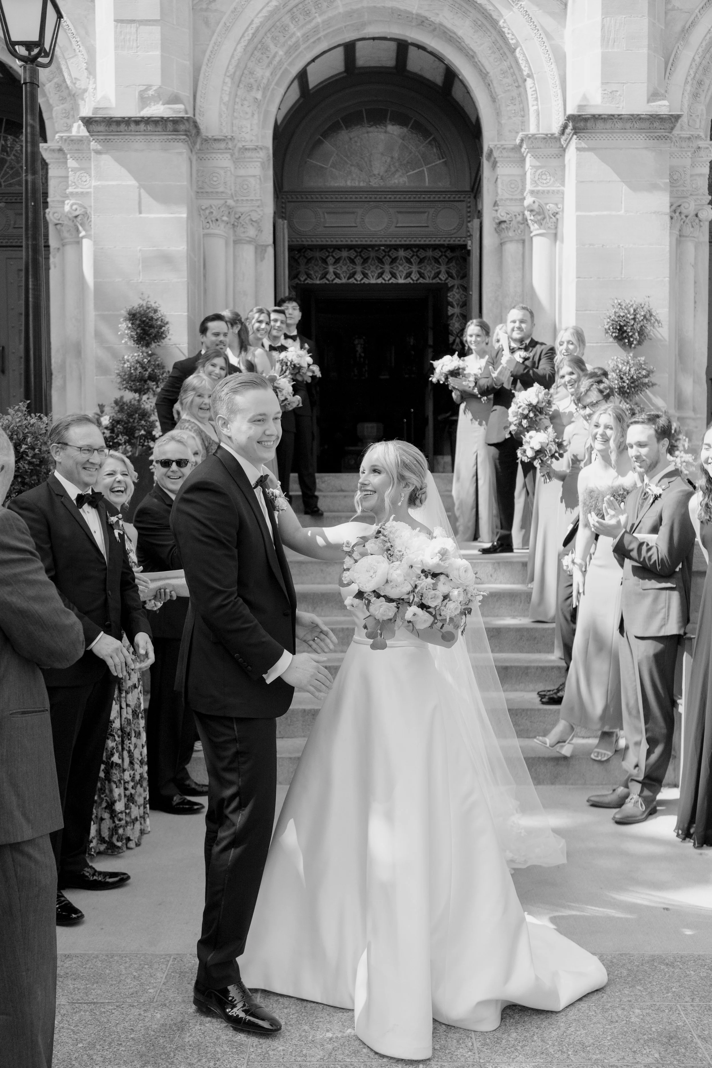 Black and white photo of a wedding ceremony outside a grand stone building, with a bride and groom holding hands and facing each other, surrounded by friends and family.