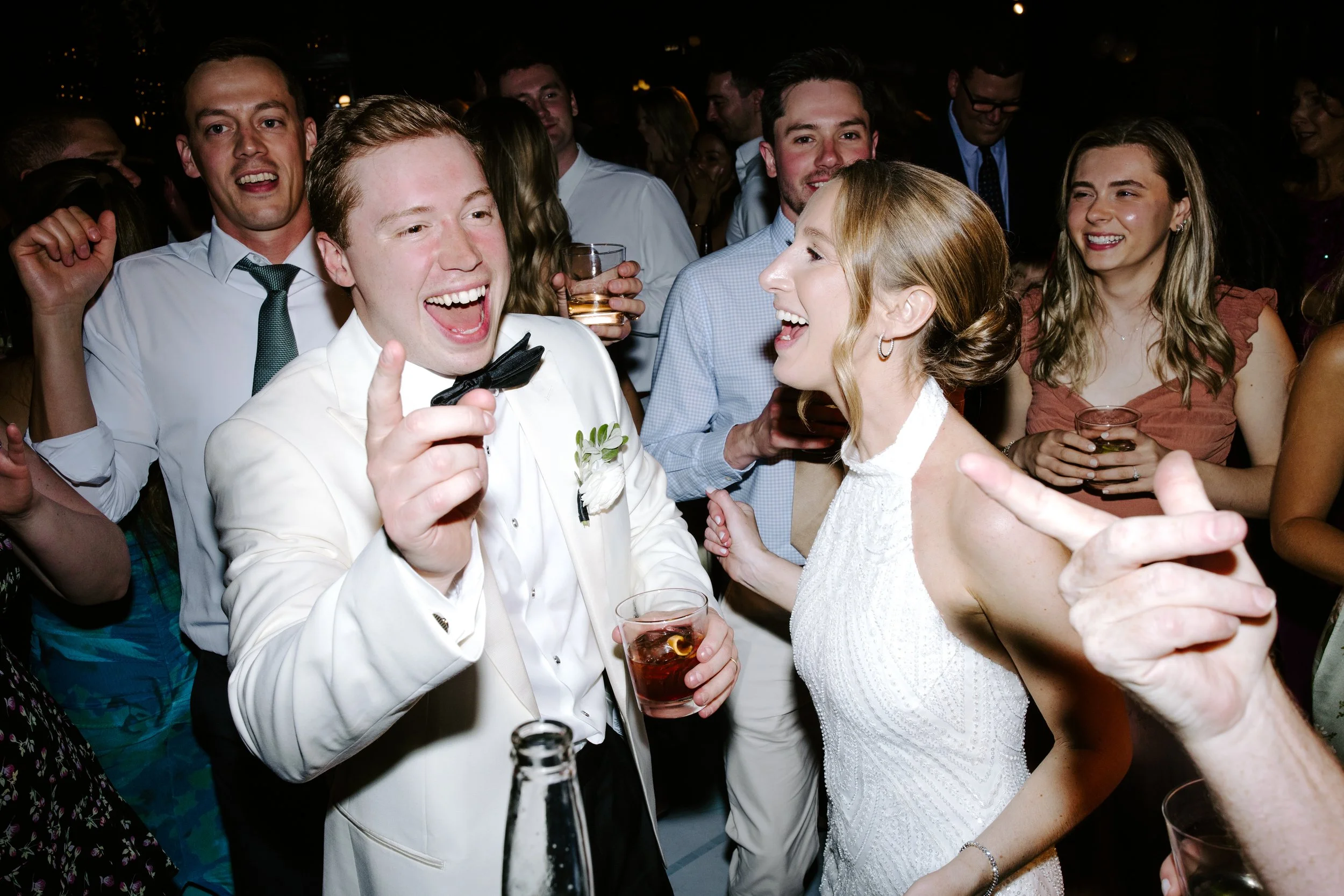 A group of people at a celebration or party, with a man in a white tuxedo with a black bow tie smiling and giving a thumbs up, and a woman in a white dress sharing a happy moment, surrounded by friends holding drinks and laughing.