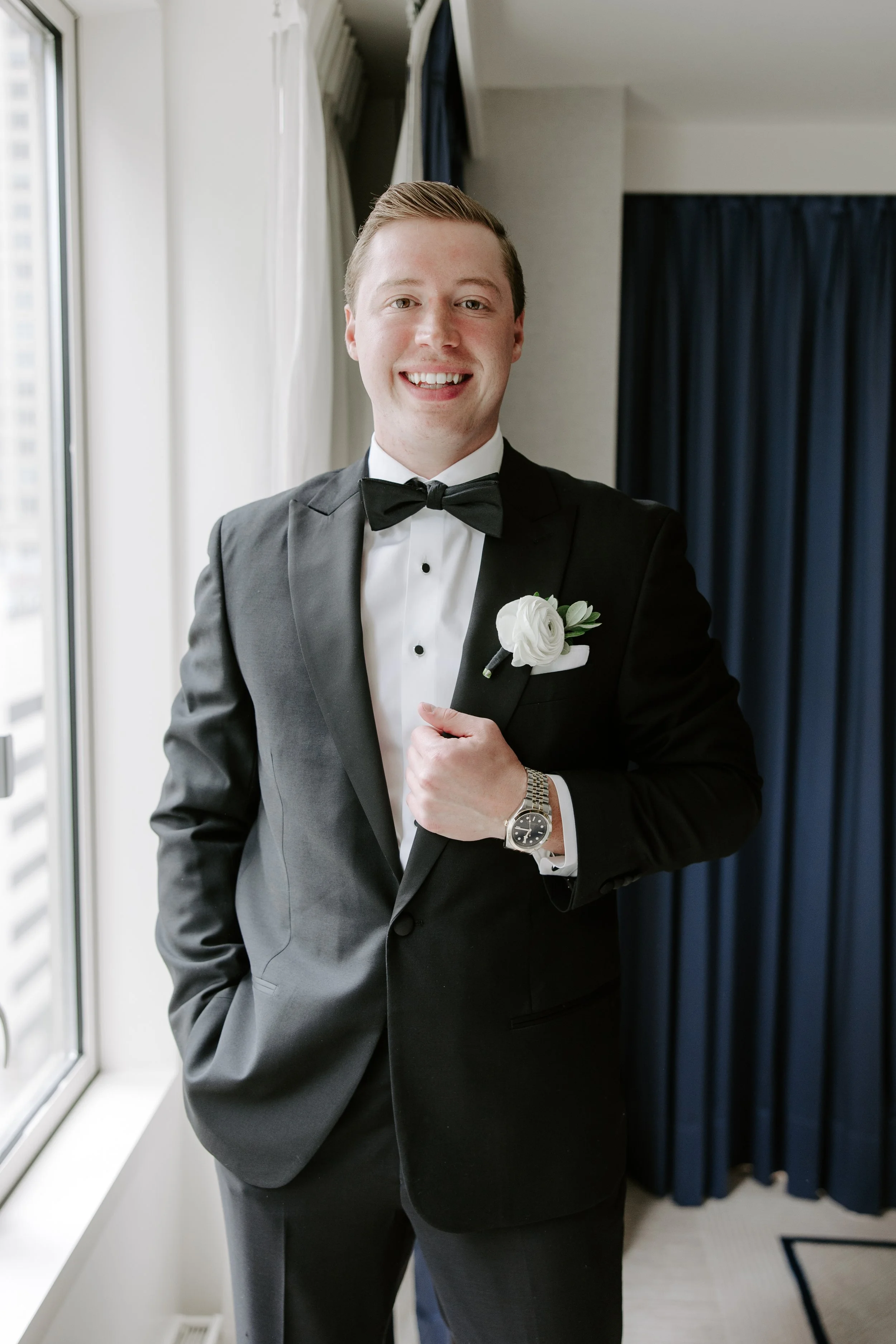 A young man dressed in a black tuxedo with a bow tie and a white boutonniere, smiling and adjusting his jacket. He is standing near a window inside a room with curtains and patterned carpet.