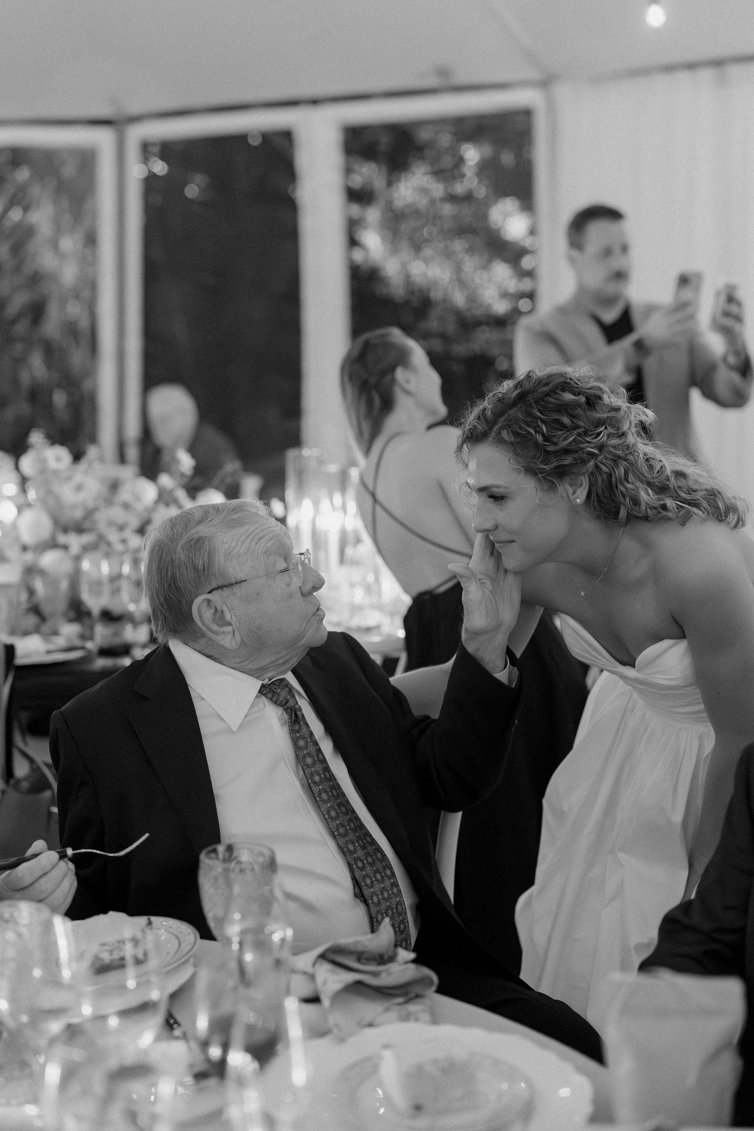 A woman in a strapless dress leaning toward an elderly man in a suit and tie at a social gathering, possibly a wedding reception, with other guests and table settings visible in the background.