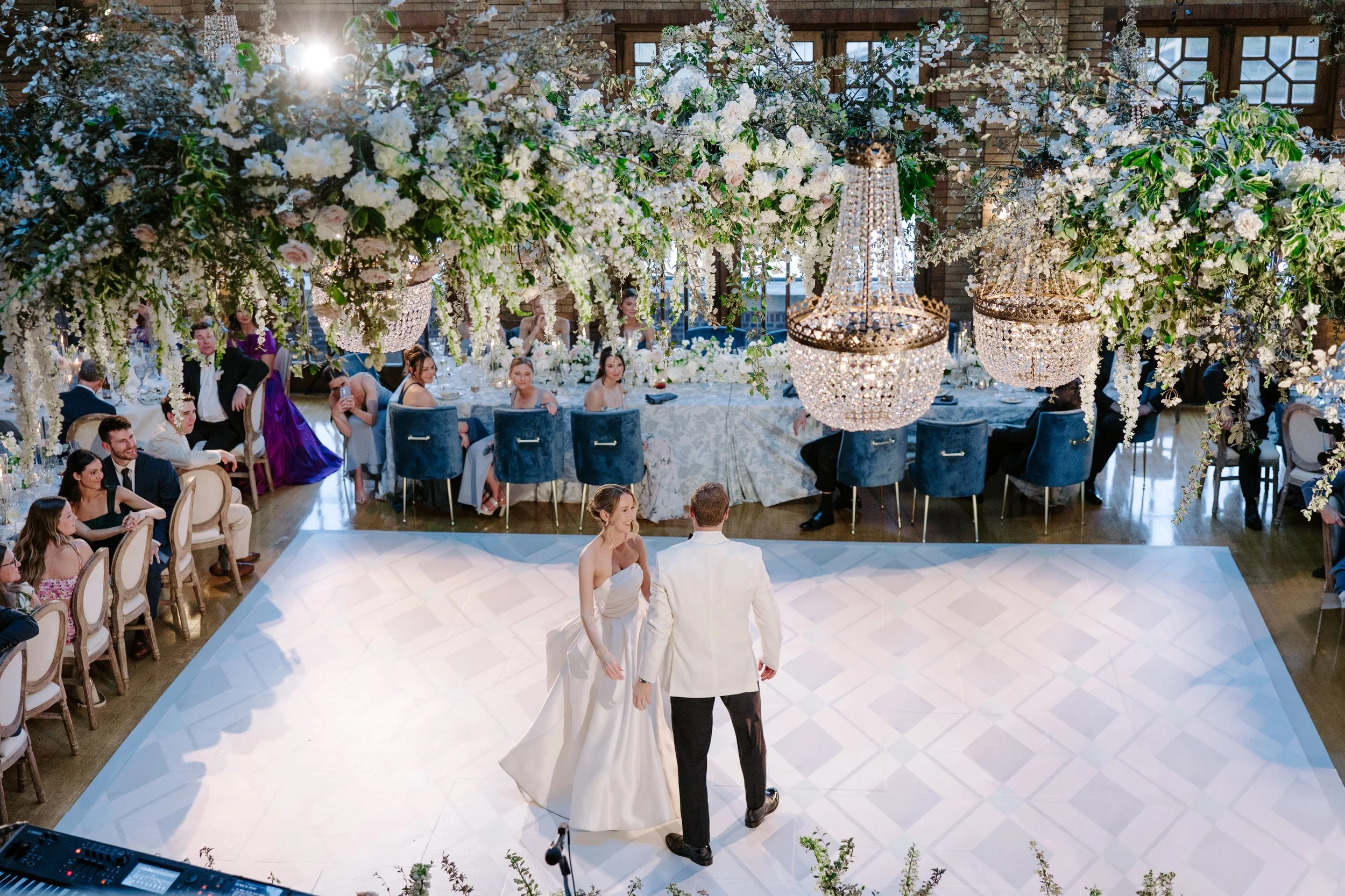 A wedding reception with a floral arch and chandeliers, featuring the bride and groom dancing on a white dance floor surrounded by seated guests.