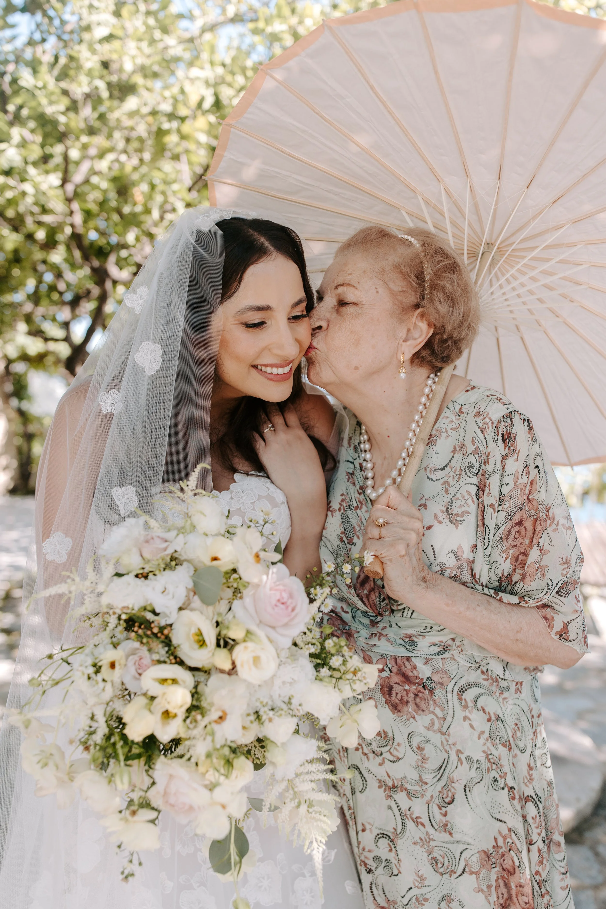 A bride wearing a lace veil and white dress is smiling as she receives a kiss on the cheek from an elderly woman holding a parasol, with a bouquet of white and pale pink flowers in the foreground. They are outdoors surrounded by green trees.