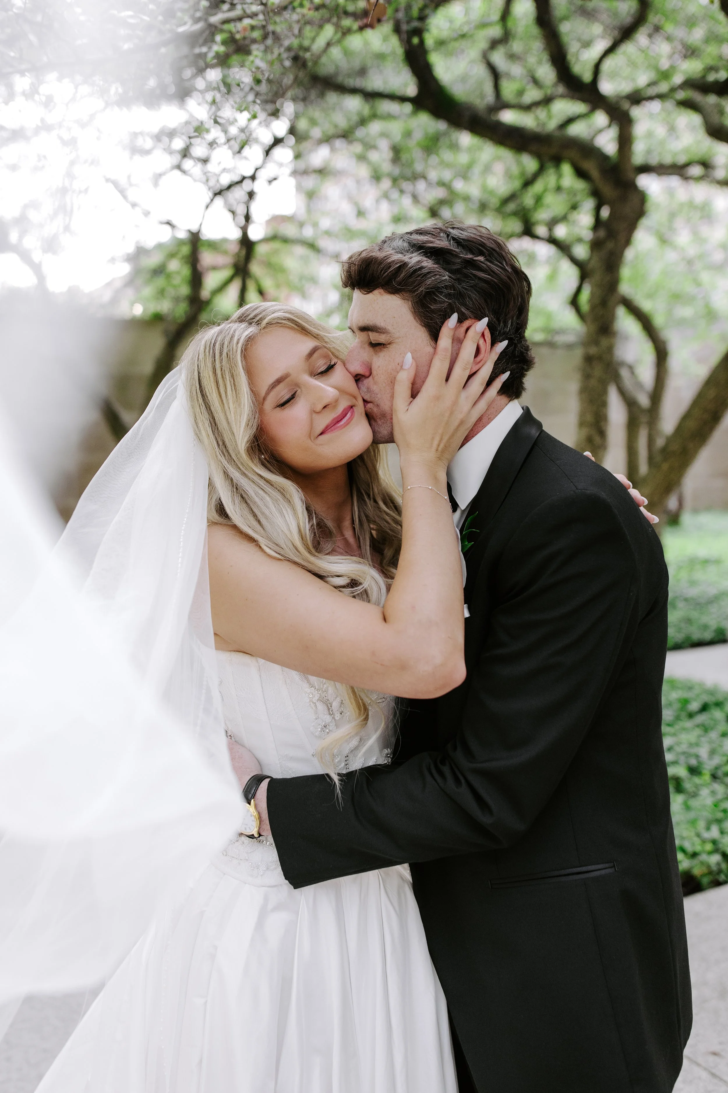 A bride and groom share a kiss outdoors, with the groom in a black tuxedo and the bride in a white wedding dress and veil, surrounded by trees.