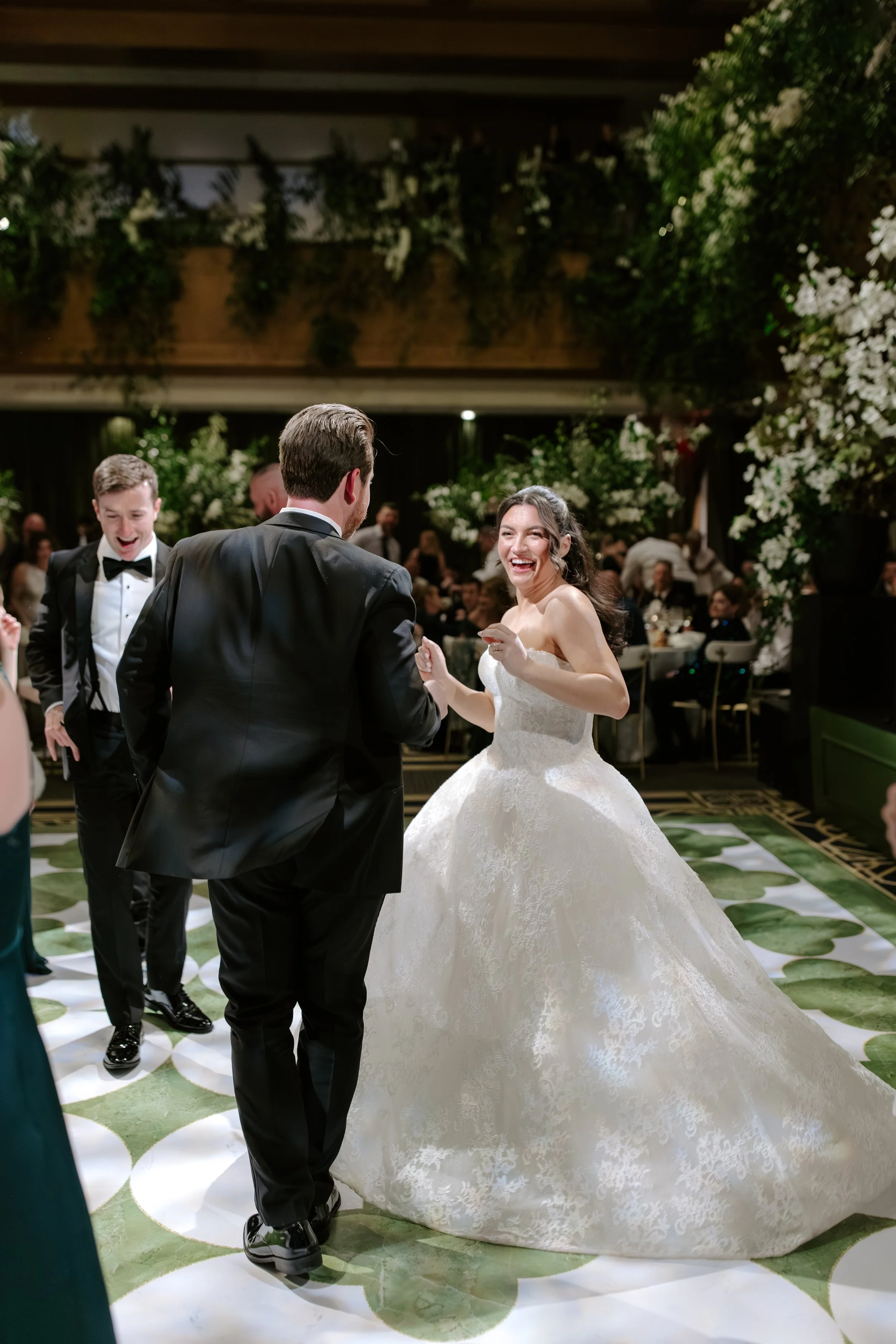 A bride and groom are dancing at their wedding reception, surrounded by guests and decorated with greenery and white flowers.