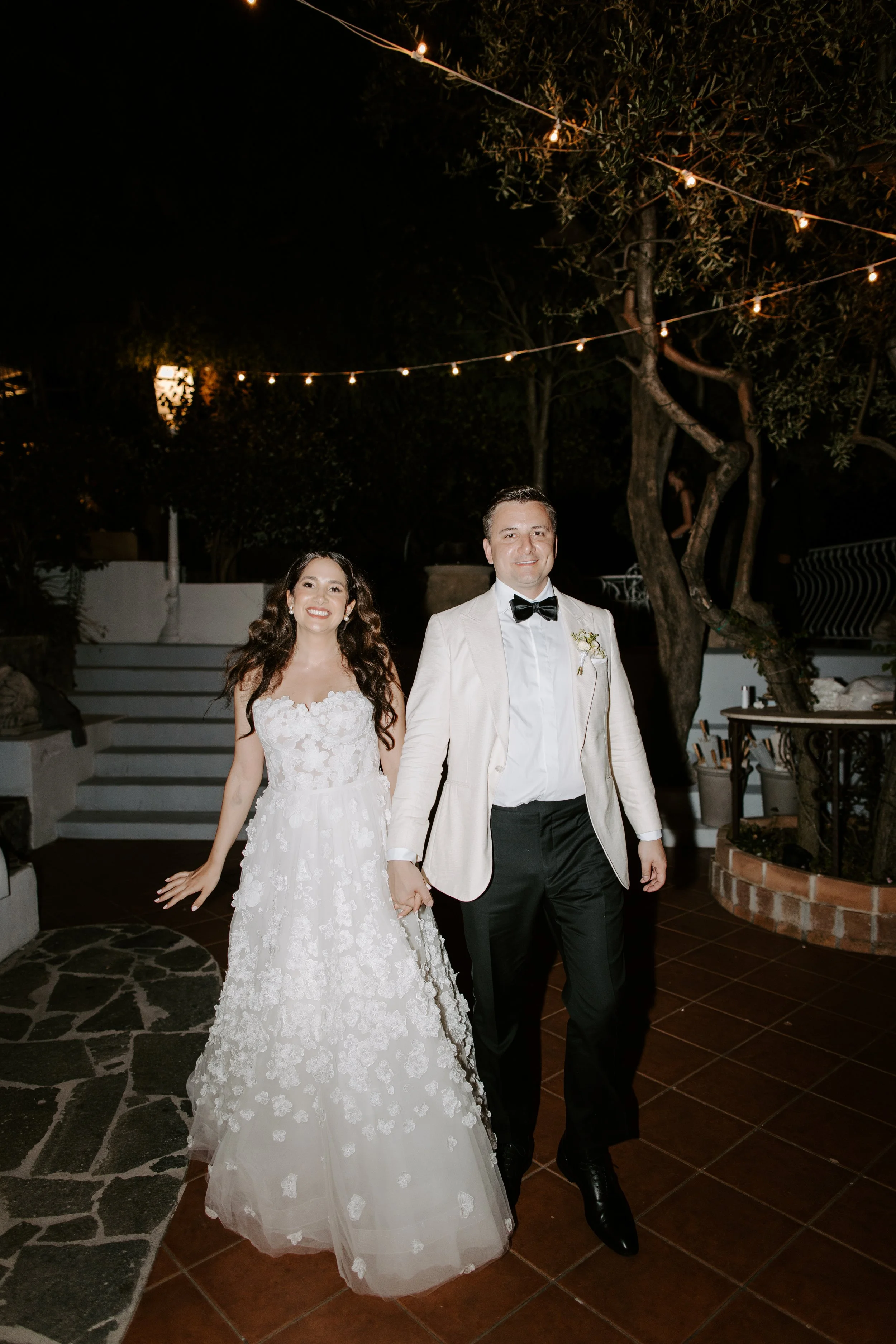 A newlywed couple walking outdoors at night, the bride in a white lace wedding dress and the groom in a tuxedo with a bow tie, holding hands and smiling.