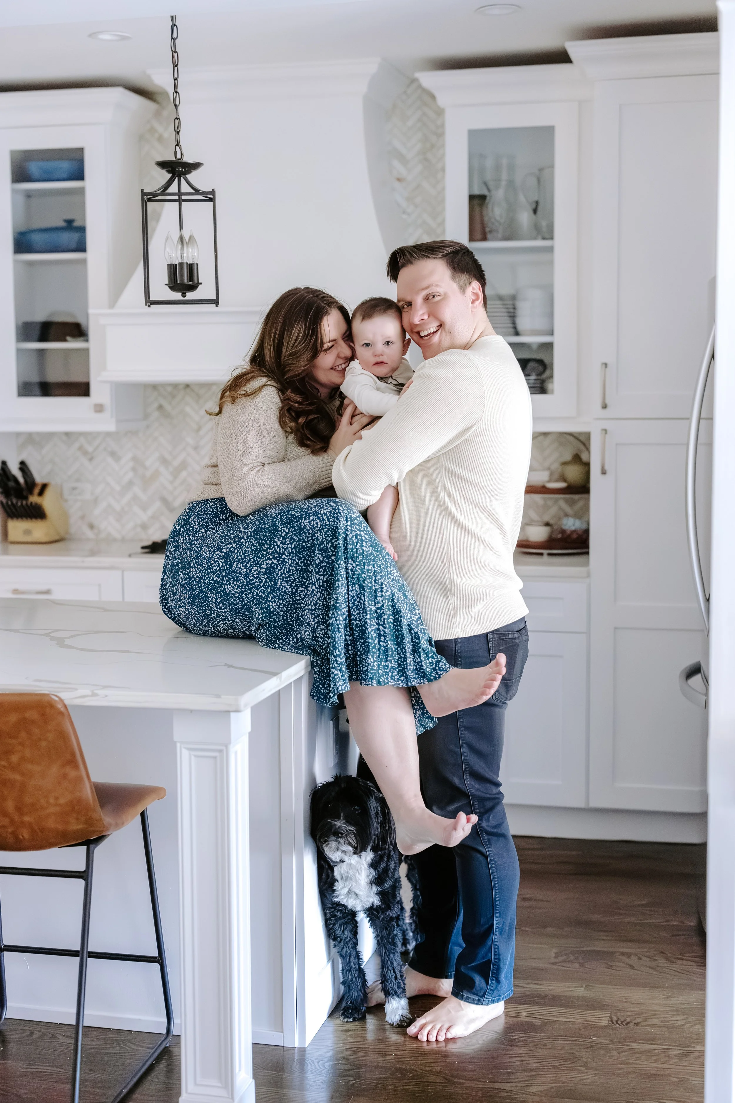 A family photo in a kitchen. The man is holding a baby, both smiling at the camera next to a woman. The woman is seated on the kitchen counter, smiling and hugging the baby. A black and white dog is standing at the woman's feet, looking up. The kitch