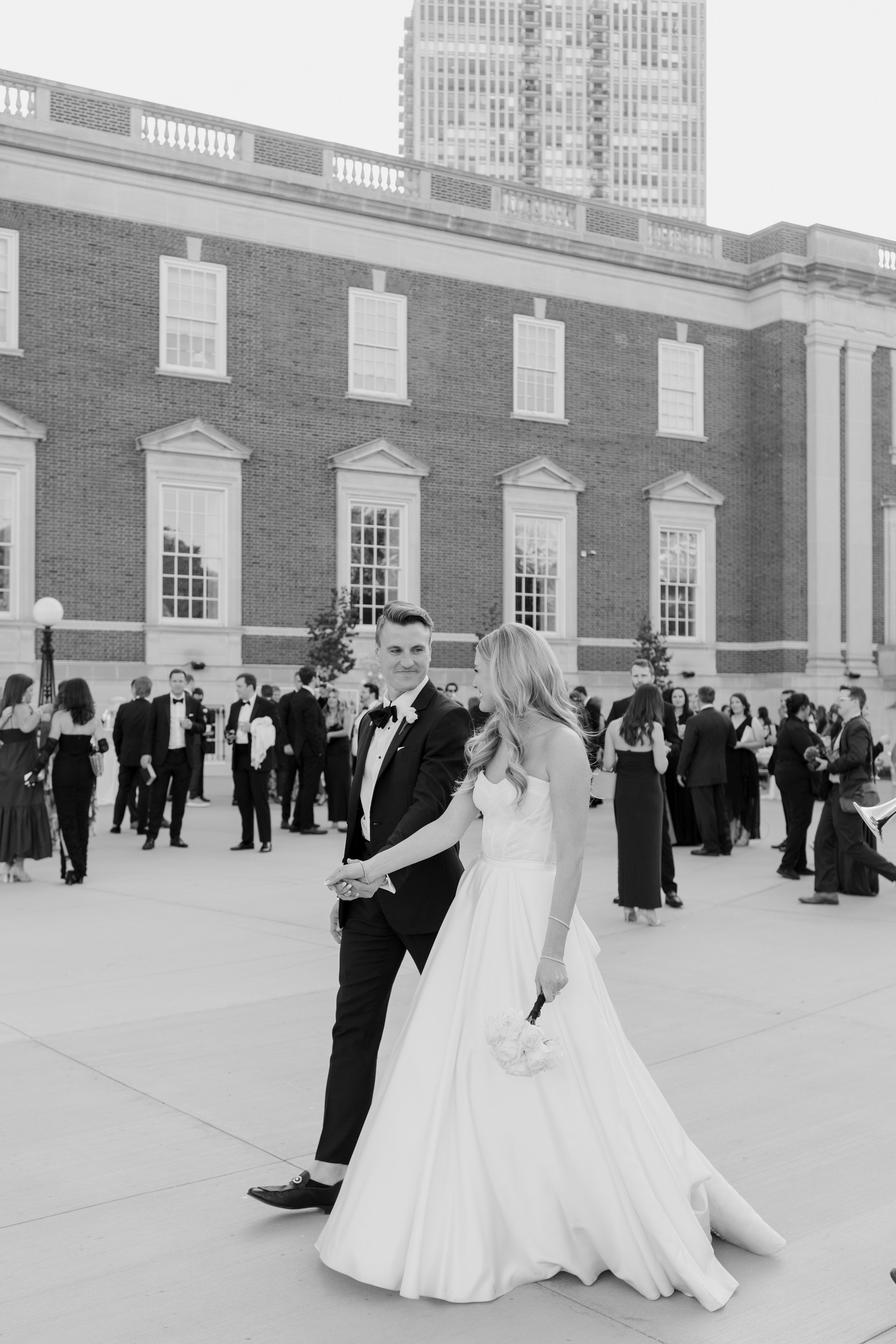 A newlywed couple walking hand in hand at a wedding reception held outdoors, surrounded by guests in formal attire, with a historic building and modern high-rise in the background.