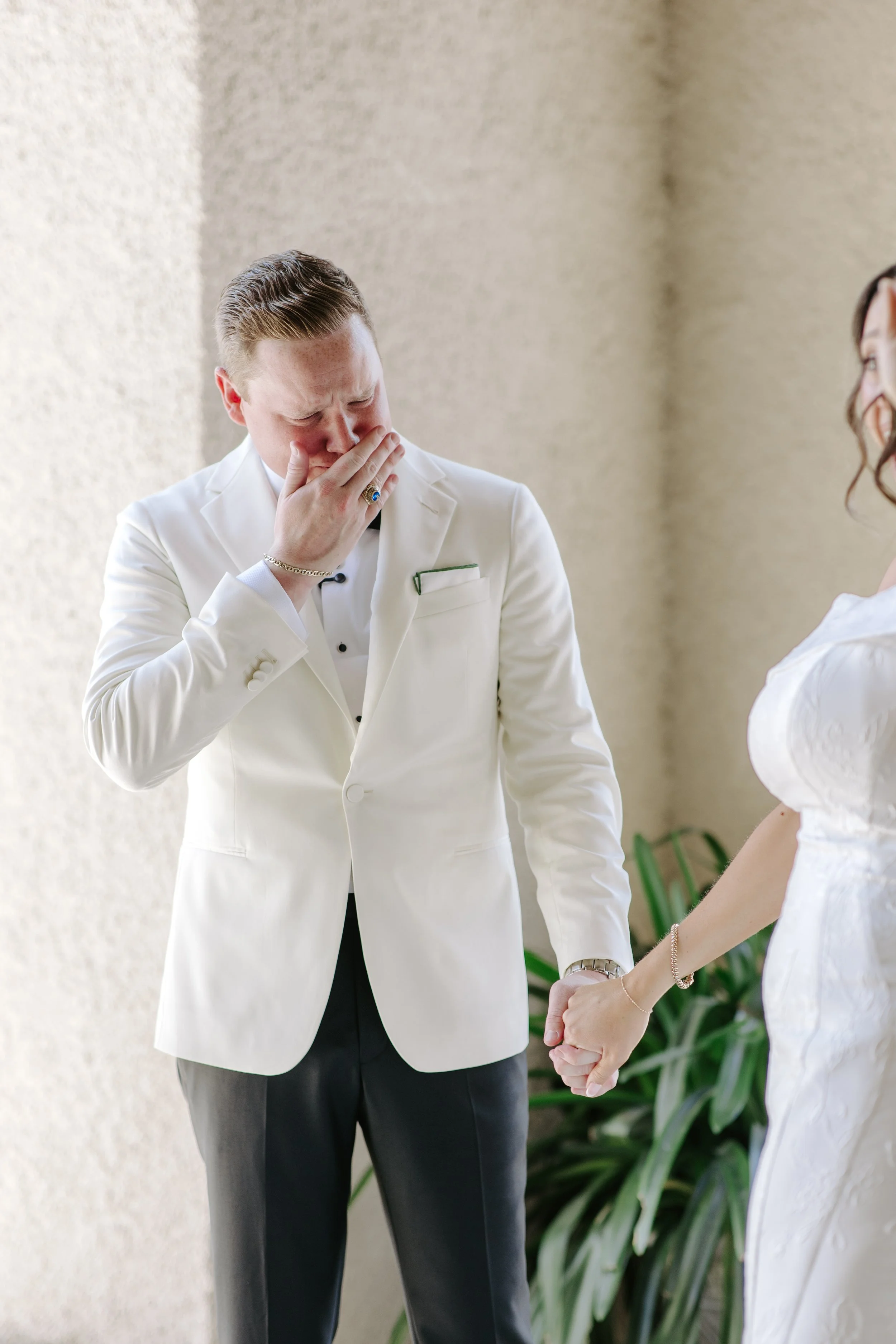 A man in a white tuxedo with black buttons and dark pants is holding a woman's hand, and they appear to be about to kiss. The man is covering his face with his right hand and has short, slicked-back hair. The woman is wearing a white dress with ruffl