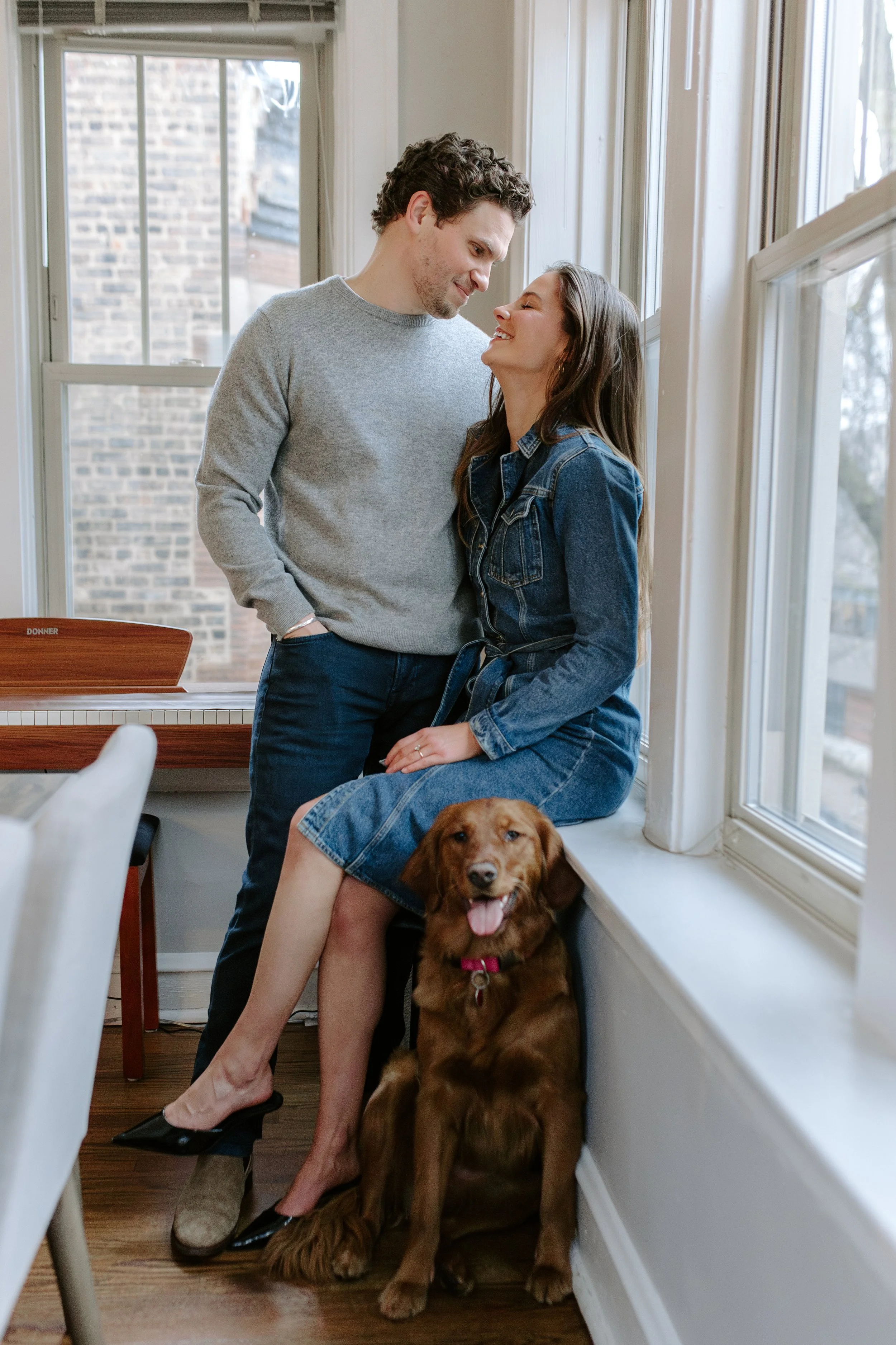 A couple with a large brown dog sitting on a windowsill in a bright room with large windows and a view of a brick building outside. The woman is sitting with her legs crossed, smiling at the man who is standing close and looking down at her. The woma