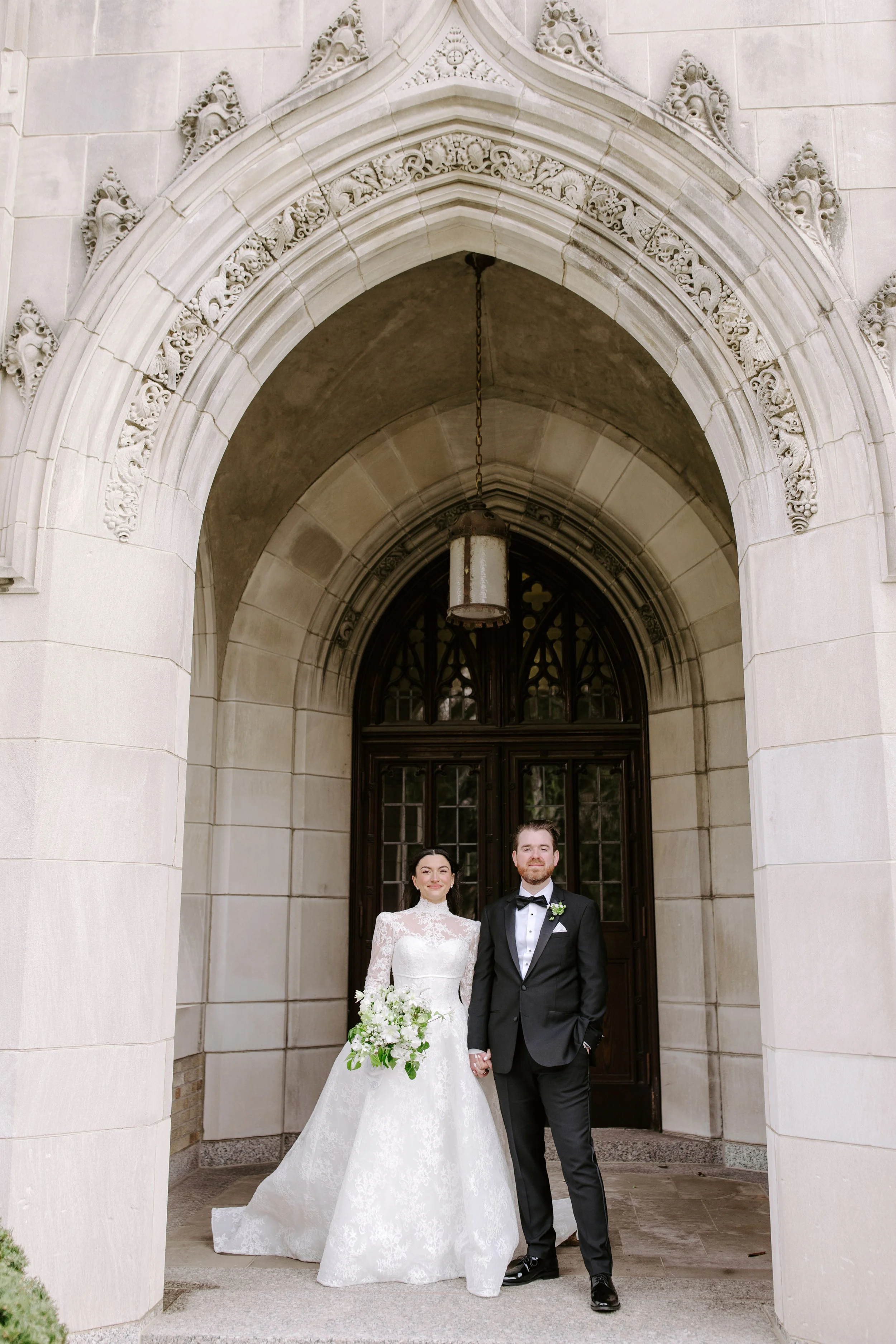 A bride and groom standing hand-in-hand in front of a historic stone building with an ornate archway and a hanging lantern. The bride wears a white lace wedding gown and holds a bouquet of white flowers with greenery. The groom wears a black tuxedo w