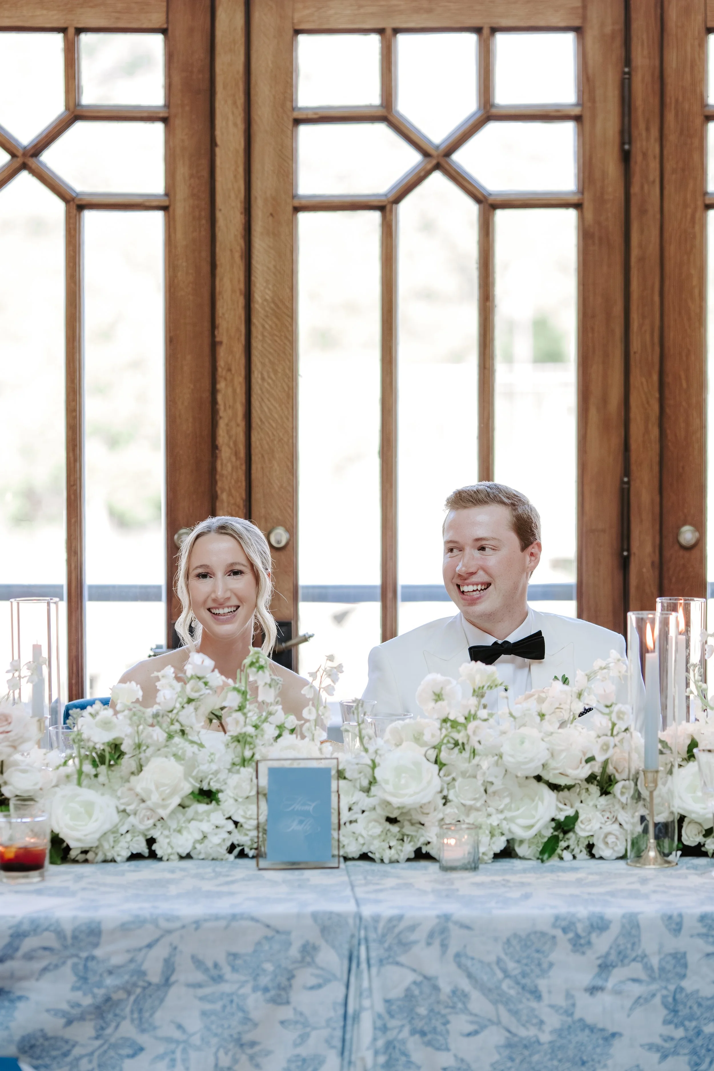 A bride and groom smiling and sitting at a decorated wedding reception table with white flowers and candles, in front of large wooden windows.