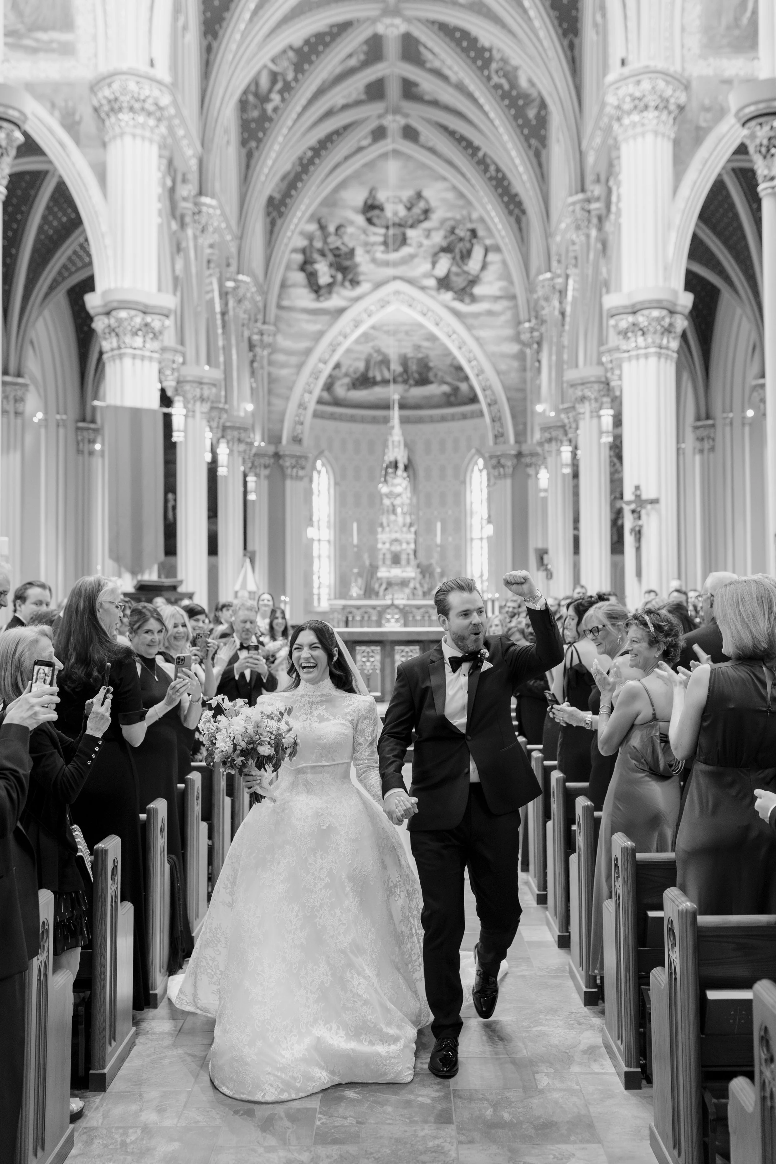 A newly married couple walking down the aisle in a church, surrounded by smiling guests taking photos and videos, with ornate columns and religious artwork in the background.